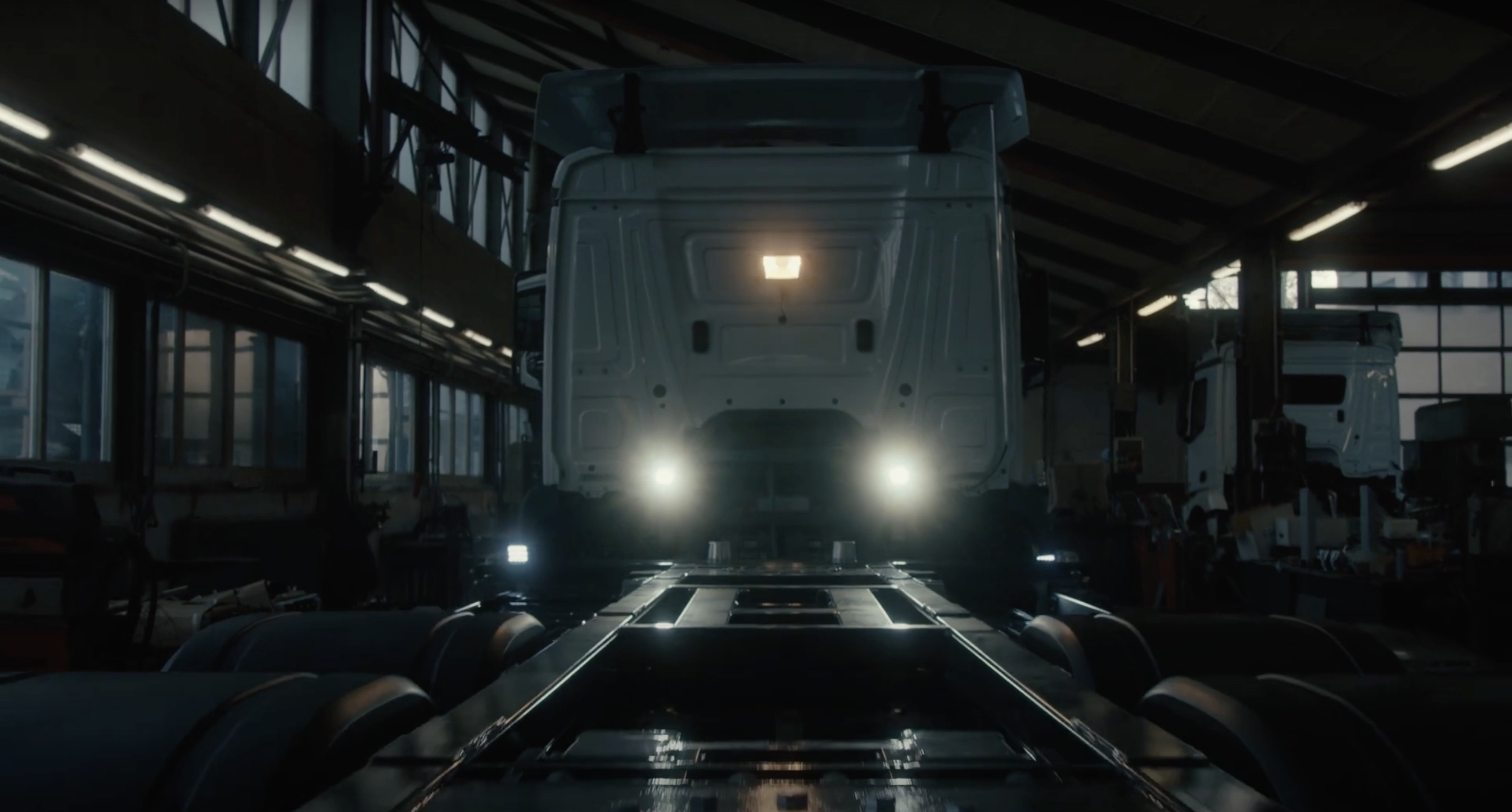 Innenansicht einer Fabrikhalle mit einem großen, weißen LKW in der Mitte, Lichtquellen an der Decke und Werkzeuge sowie Bauteile auf den Seiten.