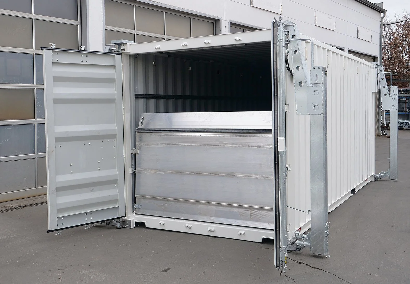 Ein leerer, versiegelter Metallcontainer mit offener Tür, auf einem asphaltierten Außenbereich neben einem modernen Gebäude.