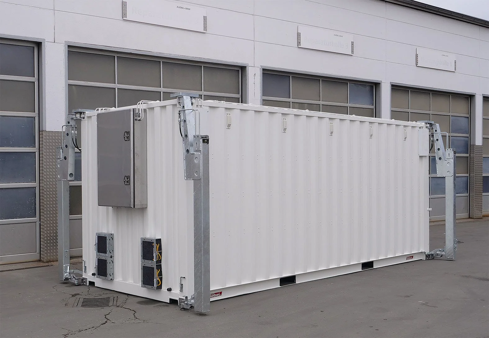 Ein weißer Kasten auf einem Parkplatz vor einer modernen Industriehalle, vermutlich ein Transformator oder Container