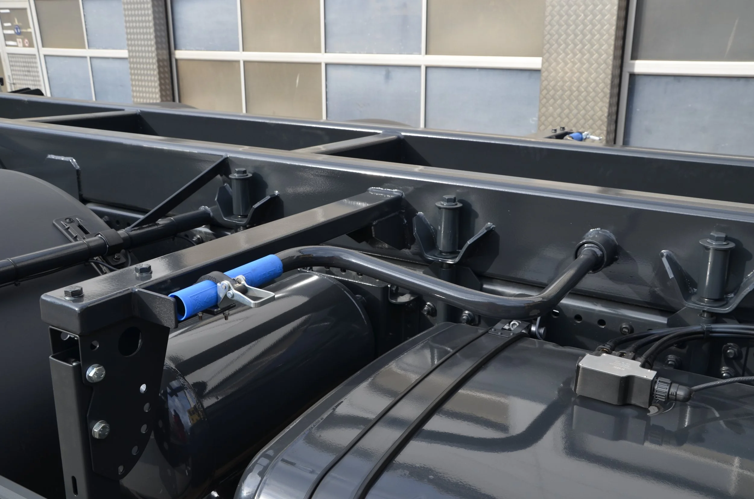 Nahaufnahme eines Fahrzeugmotors mit sichtbaren Hydraulikzylindern, Schrauben und Kunststoffrohren, im Hintergrund eine metallische Wand mit Fenstern.