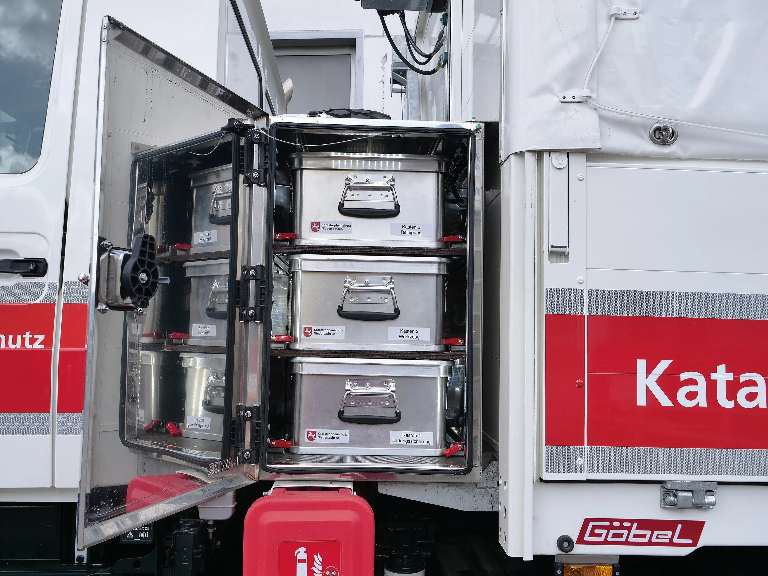 Der Bild zeigt die offene Seite eines Hydrantenwagens mit mehreren schwarzen Behältern, die mit weißen Etiketten versehen sind, auf denen die Bezeichnungen für Katastrophenschutz und Werkzeug stehen. Darunter befinden sich zwei roten Container, vermutlich für Feuerwehr- oder Gefahrgut. Der Wagen ist an einer weißen Einsatzfahrzeugseite angebracht, die eine rote Schablone mit weißen Buchstaben hat.