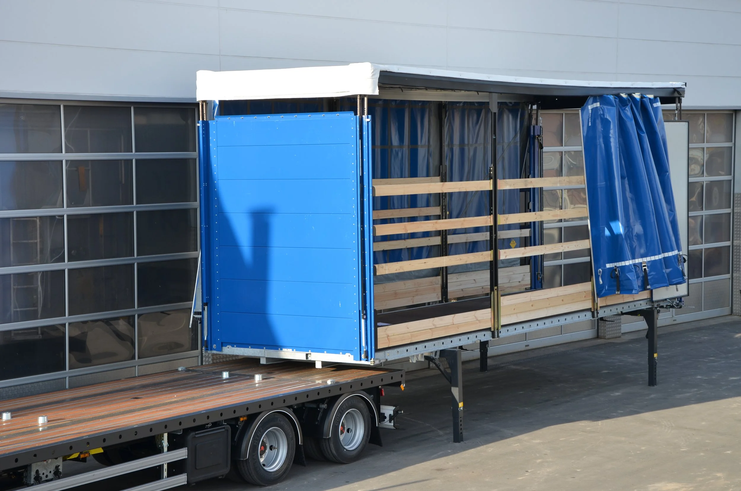 Ein Ladefahrzeug mit einem offenen, blauen Container, der auf einem Holzdecks aufgebaut ist, vor einem modernen Gebäude mit großen Fenstern.