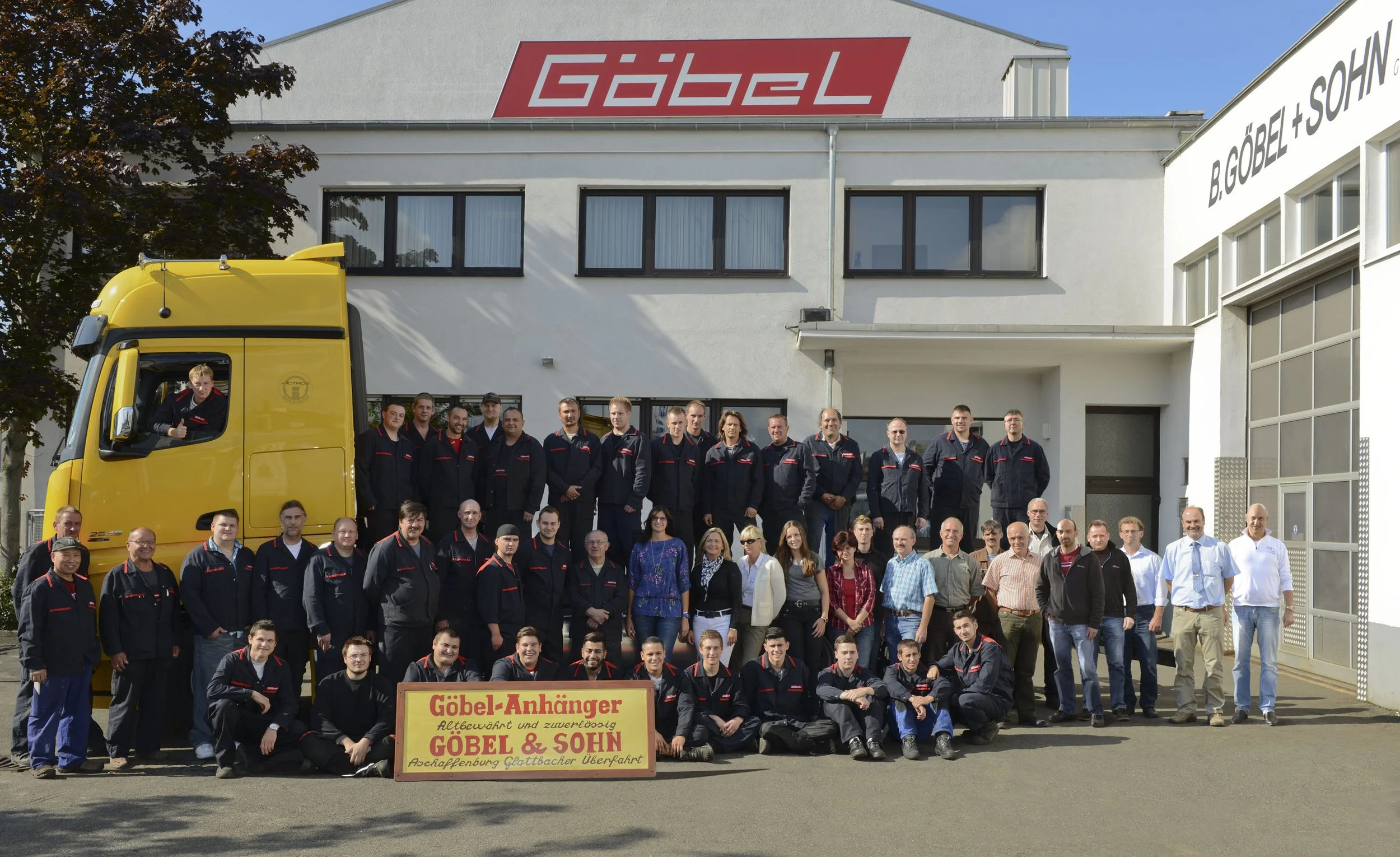 Gruppenfoto mit Mitarbeitern und Führungskräften vor dem Gebäude der Göbel Firma, das Firmenlogo ist sichtbar auf dem Dach. Es gibt eine gelbe LKW mit einem Fahrer, der grüßt. Ein Schild auf dem Boden bewirbt Göbel-Anhänger.