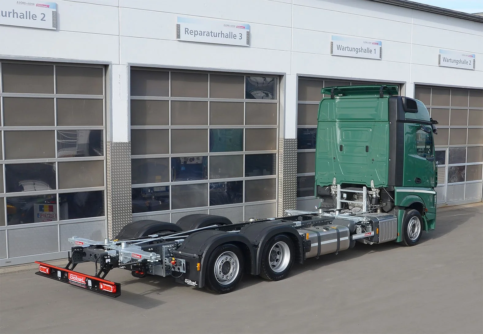 Ein grüner LKW ohne Auflieger vor einer weißen Industriehalle mit mehreren Garagentoren. Die Halle hat Schilder mit Bezeichnungen wie 'Reparaturhalle 3' und 'Wartungshalle 1'.