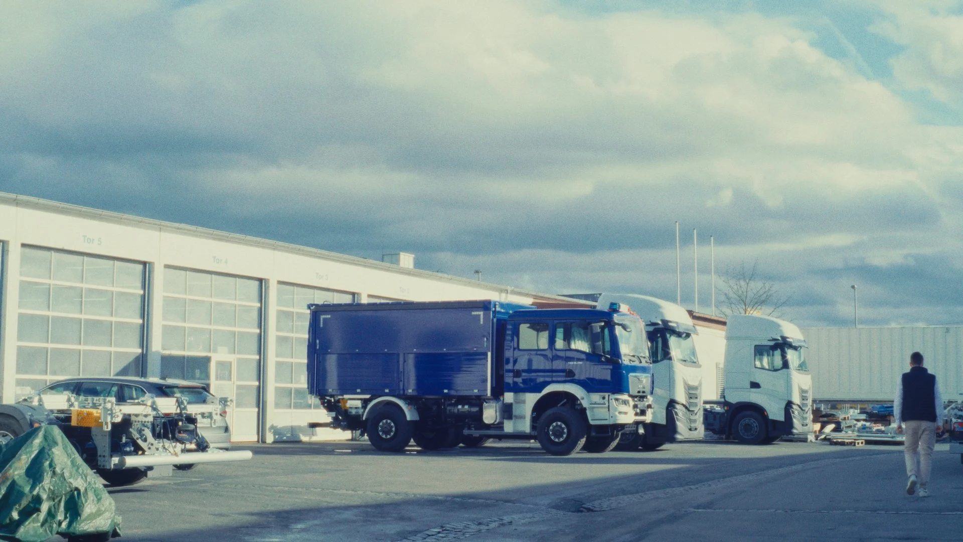 LKW und Autos auf einem Parkplatz vor einem Gebäude mit großen Fenstern und Wolken am Himmel, eine Person läuft vorbei.