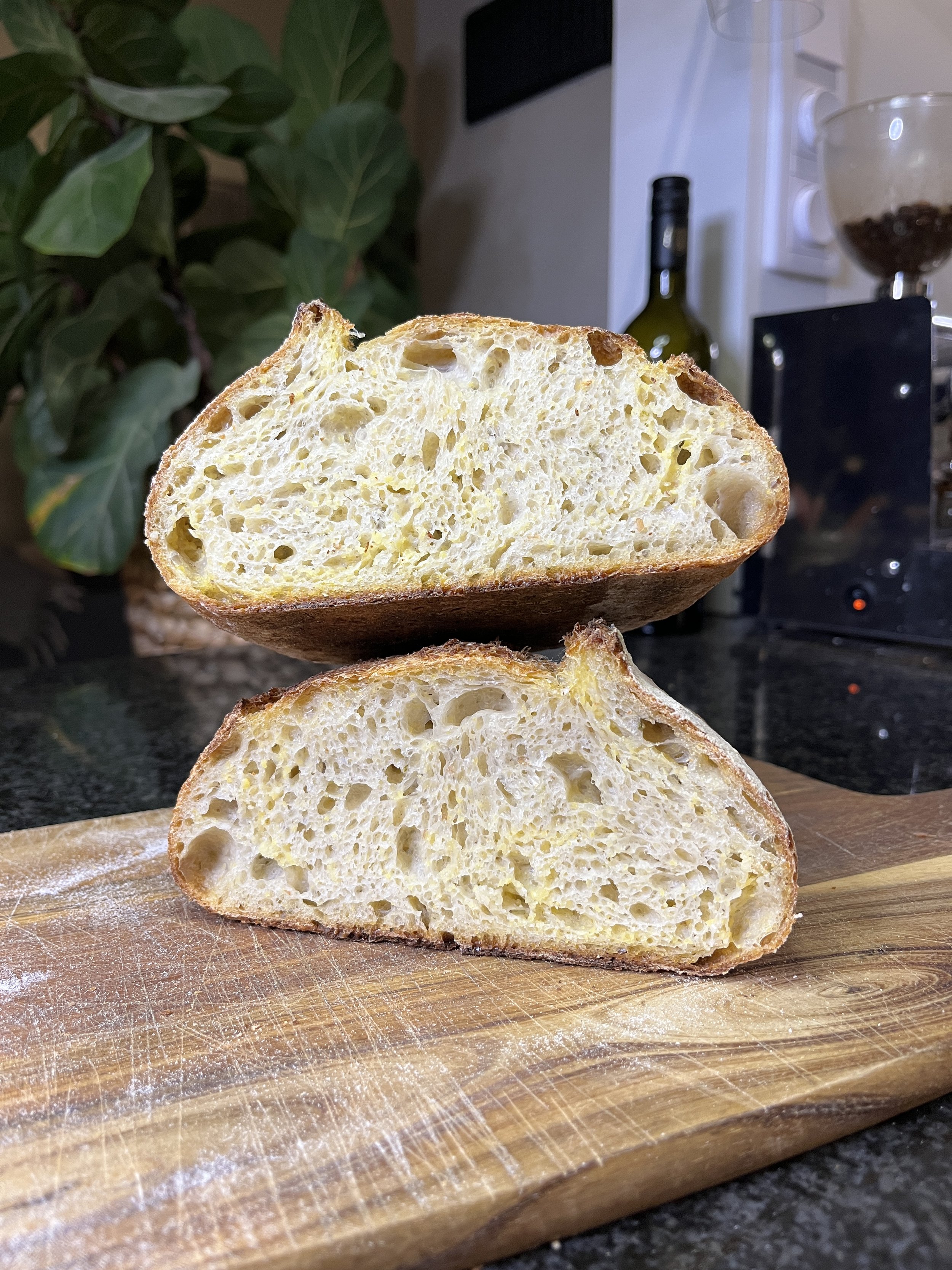 Två halvor av ett nybakat, luftigt bröd på en träskärbräda i ett kök, bakgrund med köksutrustning och växt.