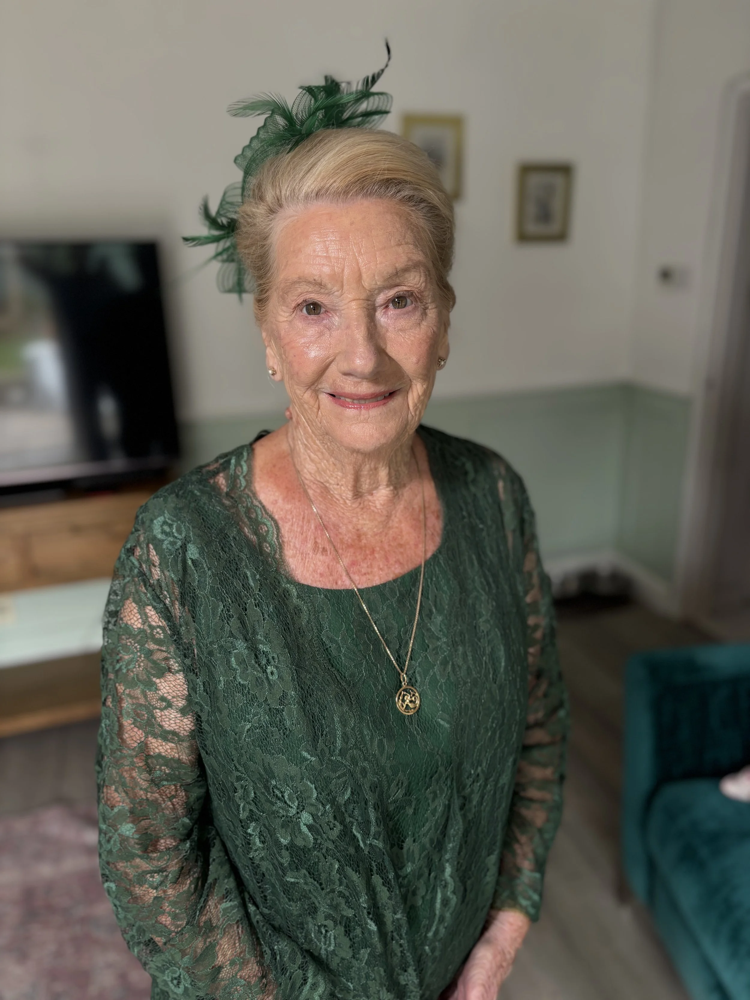 Older woman with gray hair styled in an updo, wearing a green hat with feathers, a green lace dress, and a gold pendant necklace, standing in a room with framed pictures on the wall.