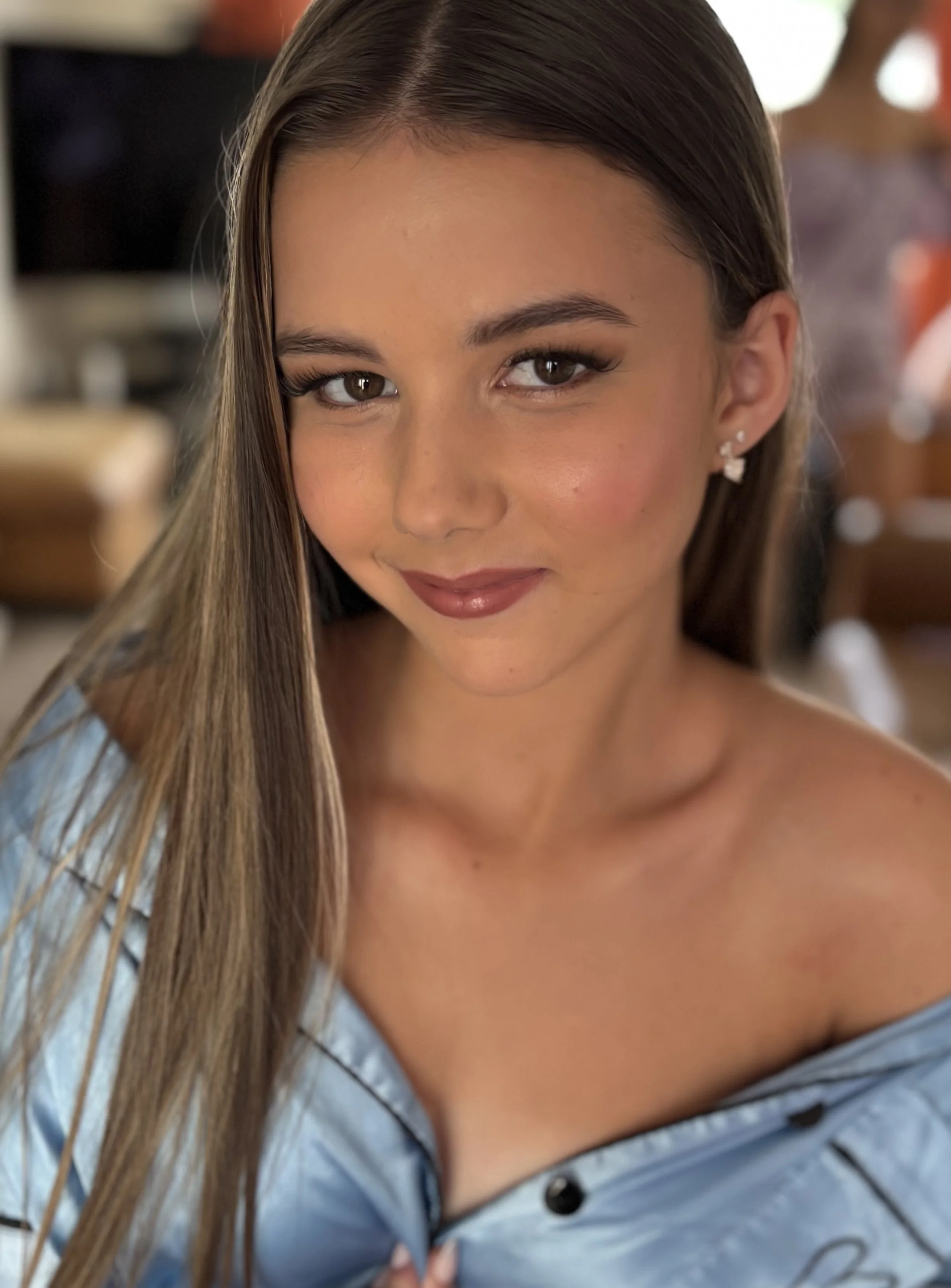 A close-up of a woman with long brown hair, light skin, and brown eyes, wearing earrings and a blue striped shirt.
