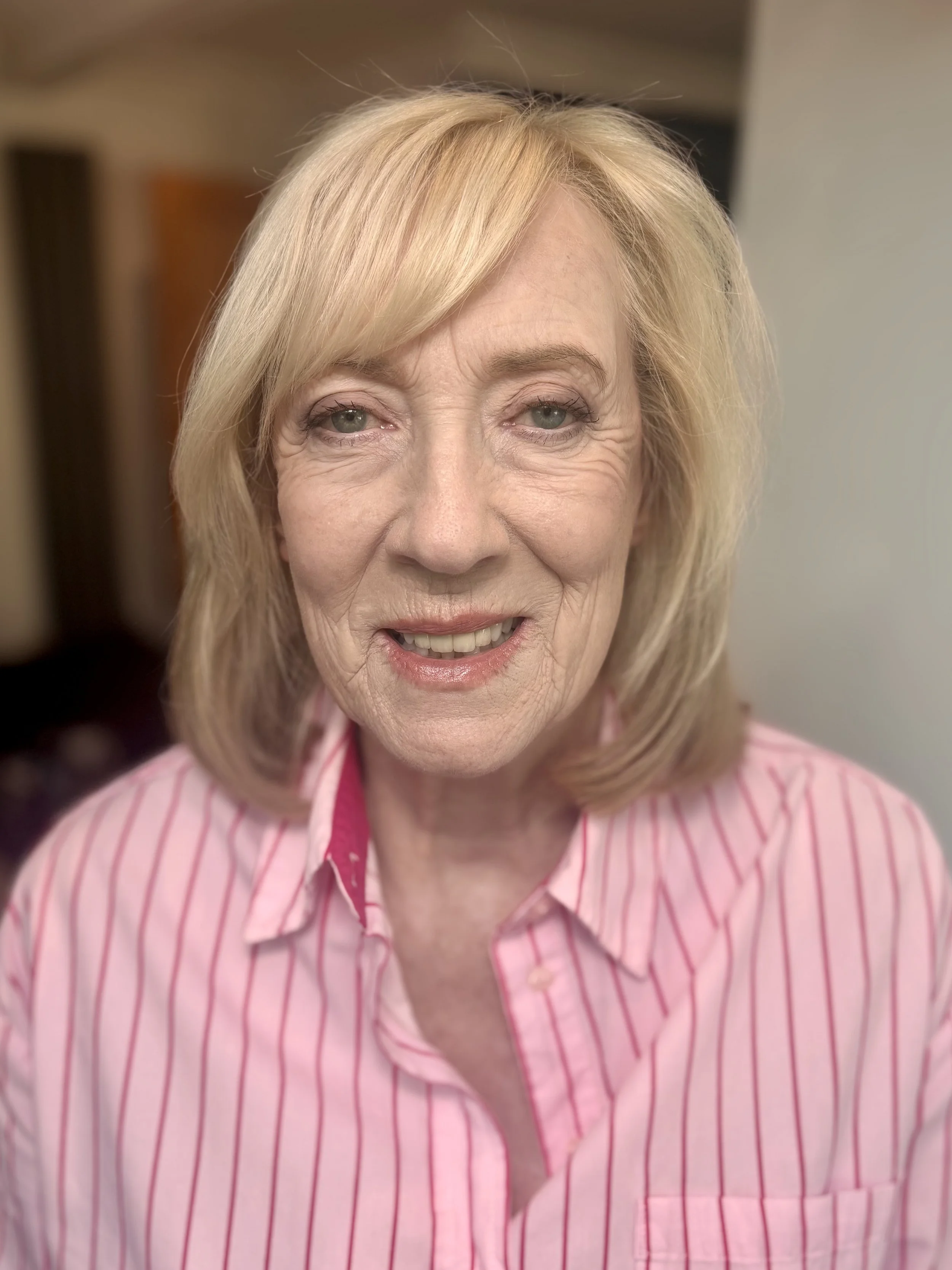 Close-up of an elderly woman with blonde hair wearing a pink and white striped shirt.