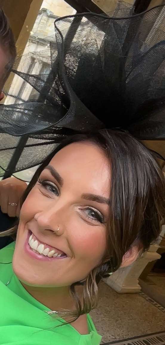 A woman with short hair smiling, wearing a neon green top, and a black fascinator hat, taking a selfie indoors with a wooden surface visible in the background.