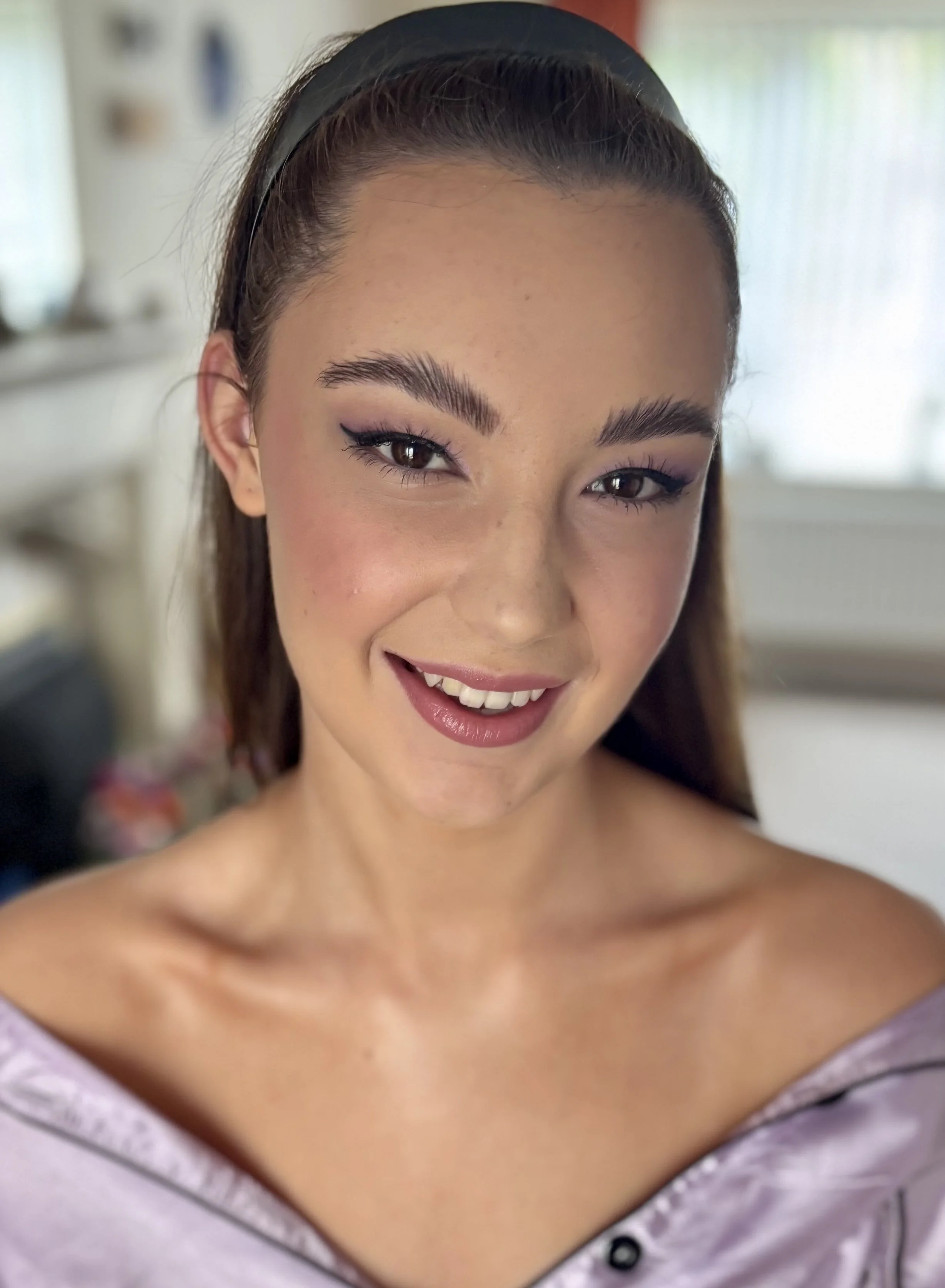 Close-up of a young woman's face smiling, showing her teeth. She has light skin, brown eyes, and long brown hair pulled back with a headband. She is wearing makeup, including eyeliner and blush.