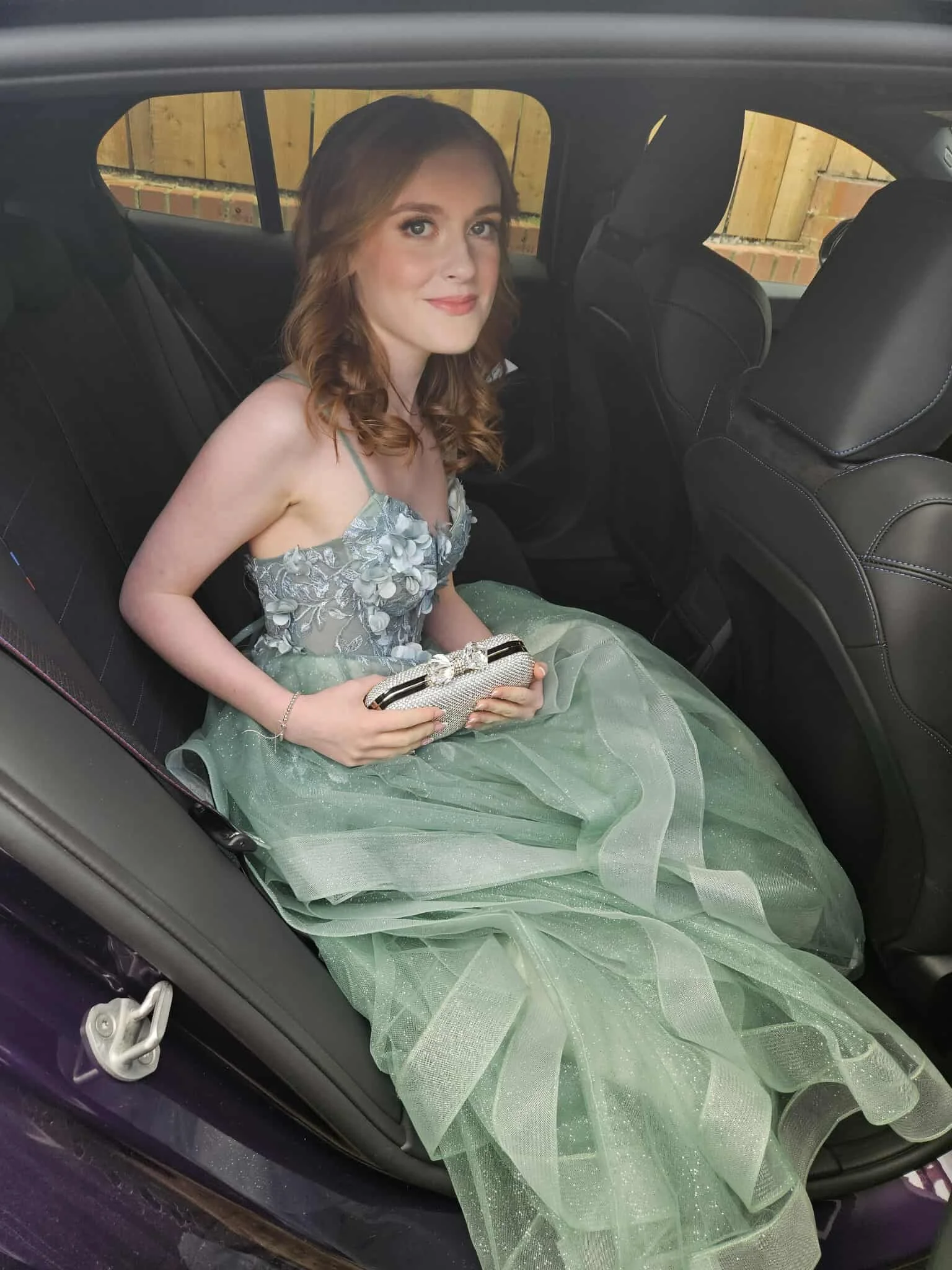 Young woman in a formal dress sitting in the back seat of a car, holding a clutch, with a wooden fence visible through the windows.