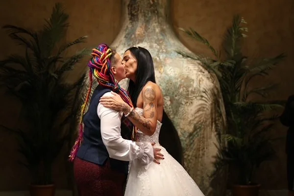 Two women in wedding attire sharing a kiss indoors, with a large decorative vase and plants in the background.