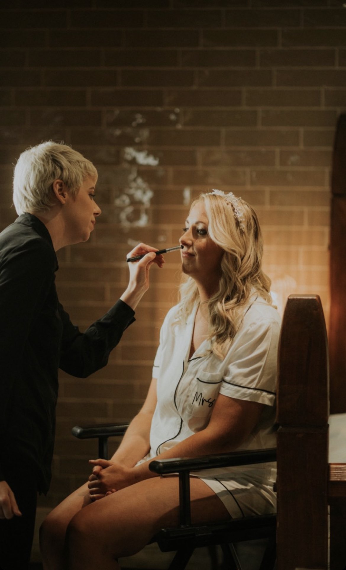 Leah Kelly, makeup artist, applying makeup to a seated woman with a tiara, wearing pajamas labeled 'Mrs', in a room with a wooden wall.