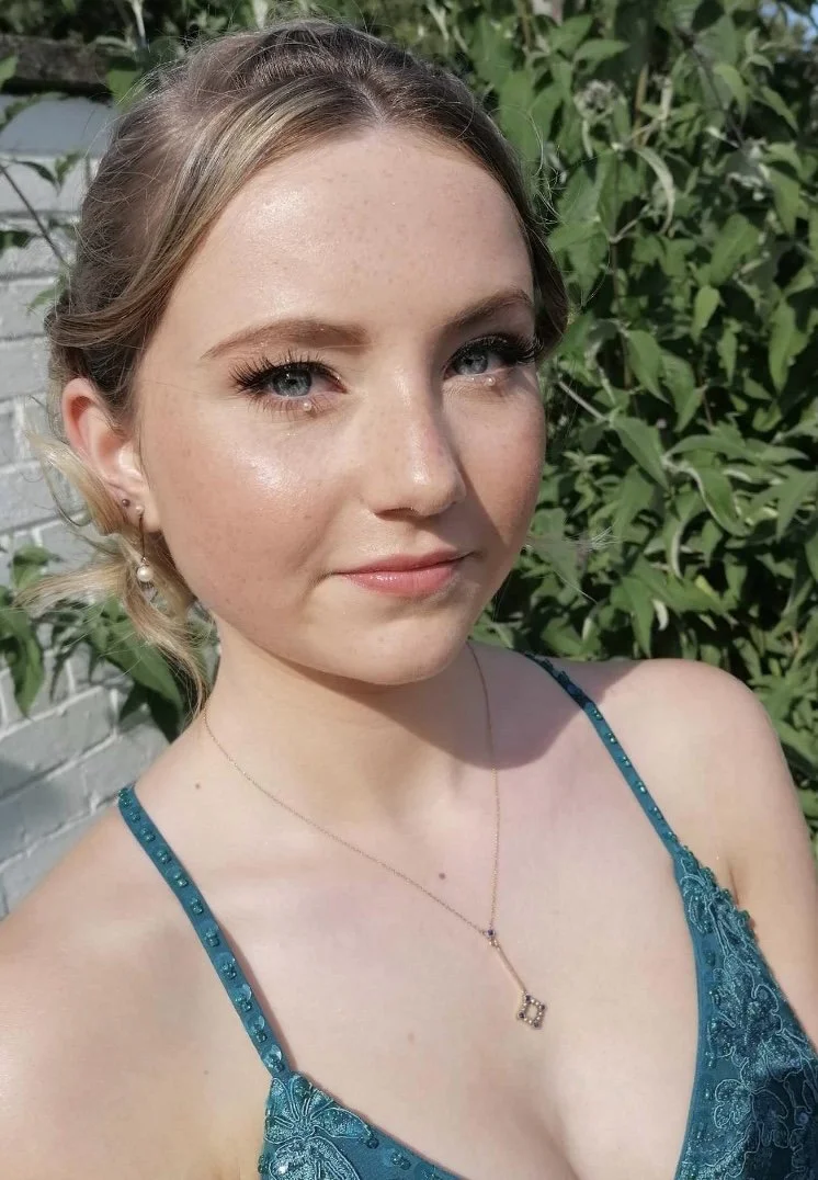A young woman with blonde hair and blue eyes taking a selfie outdoors, wearing a teal dress with thin straps and a necklace, with greenery and a white brick wall in the background.