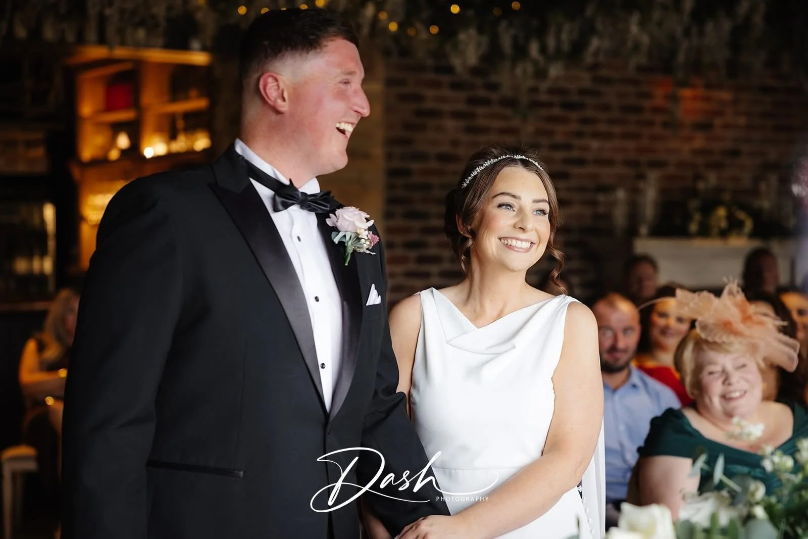 A smiling bride and groom during their wedding ceremony, with wedding guests in the background, inside a warmly lit venue with brick walls.