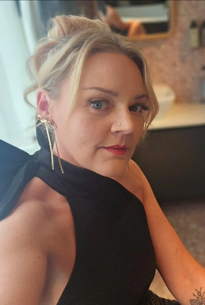 A woman with blonde hair in an updo, wearing a black sleeveless top, gold hoop earrings, and a nose ring, sitting in a cafe or restaurant.
