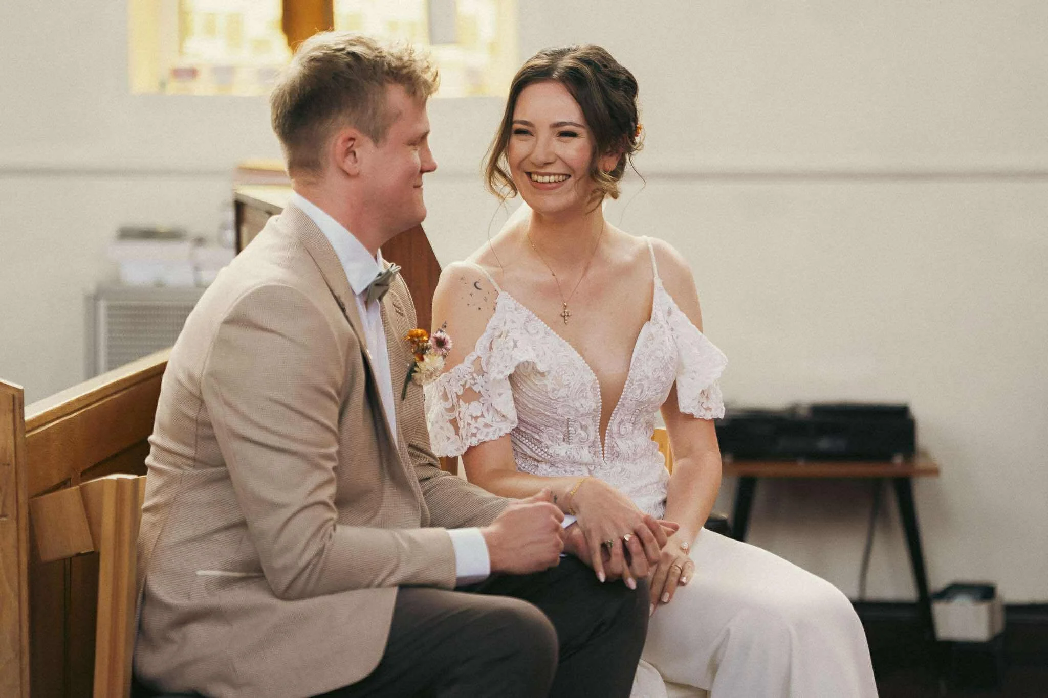 A smiling woman in a white lace dress sits next to a man in a beige suit with a boutonniere, both seated on a wooden pew inside a church or chapel, holding hands.