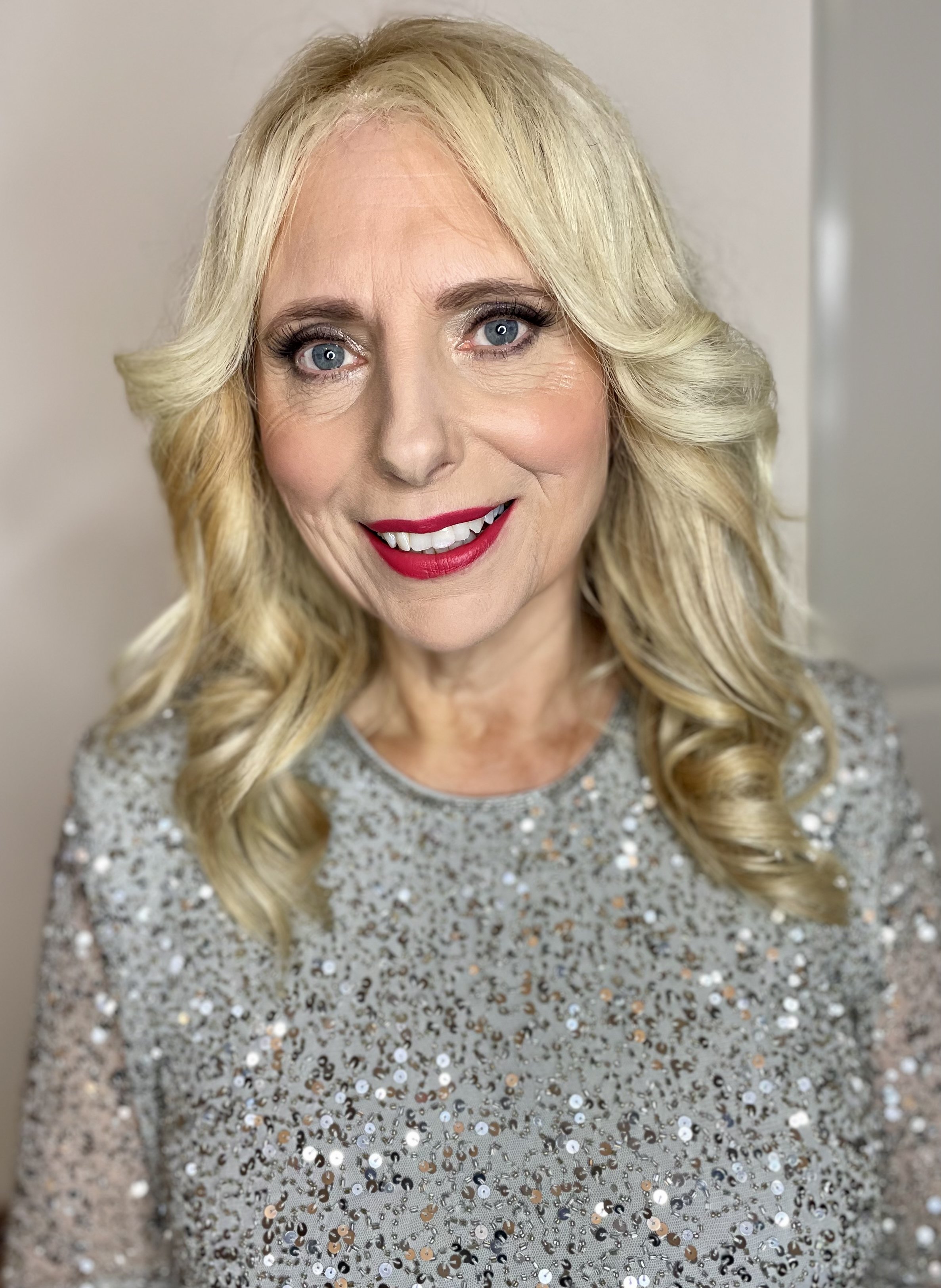 A woman with blonde, wavy hair and bright blue eyes wearing a sequined silver dress and red lipstick smiling at the camera.