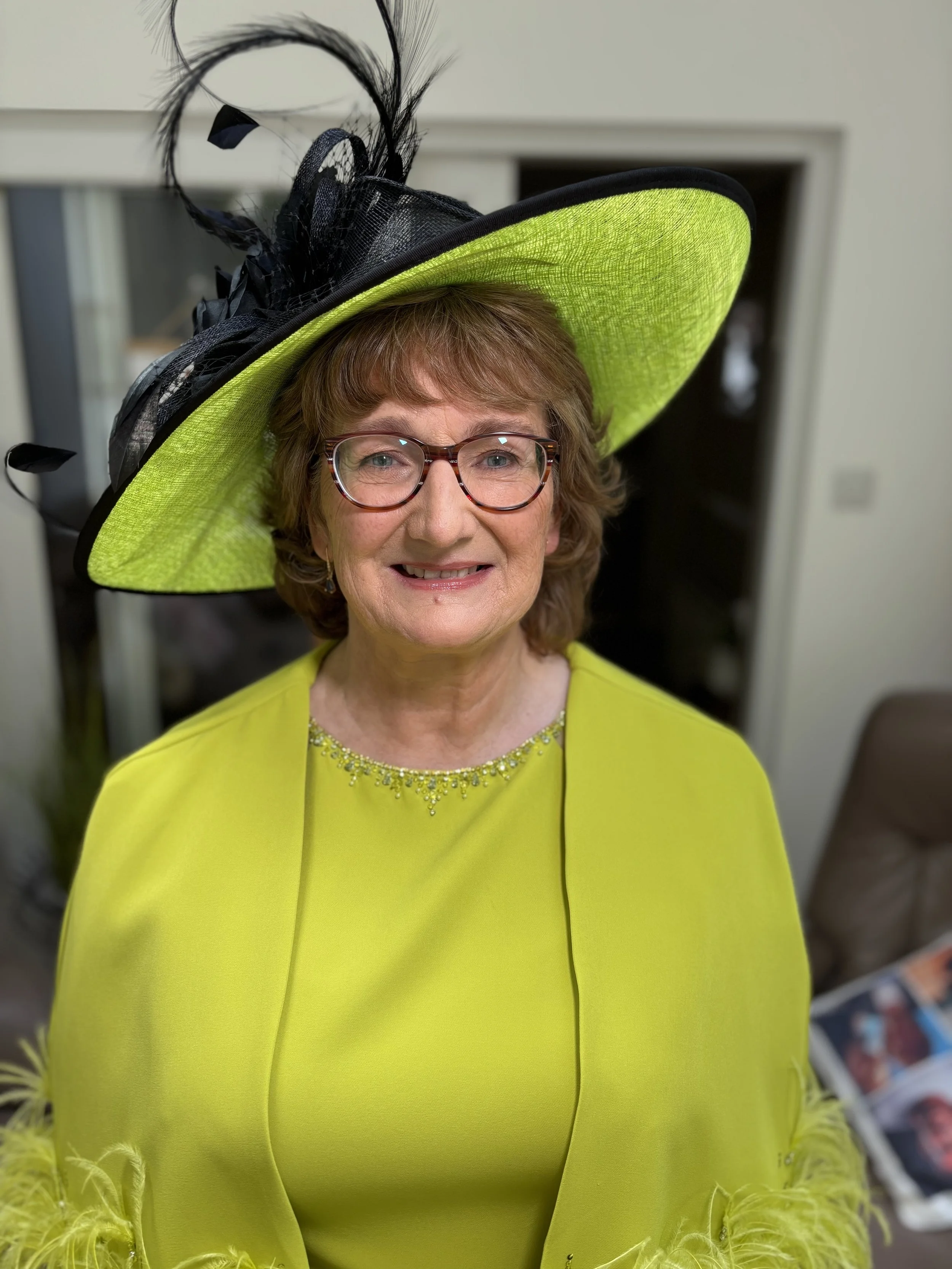 A woman wearing a bright yellow dress and a large matching yellow hat with black feather decorations and black netting, smiling at the camera.