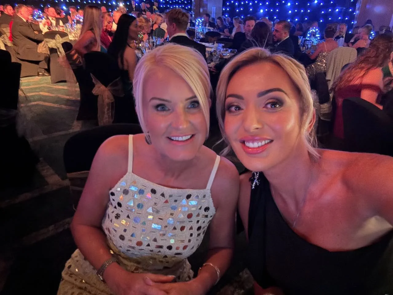 Two women taking a selfie at a formal event with many attendees, blue string lights, and decorated tables in the background.