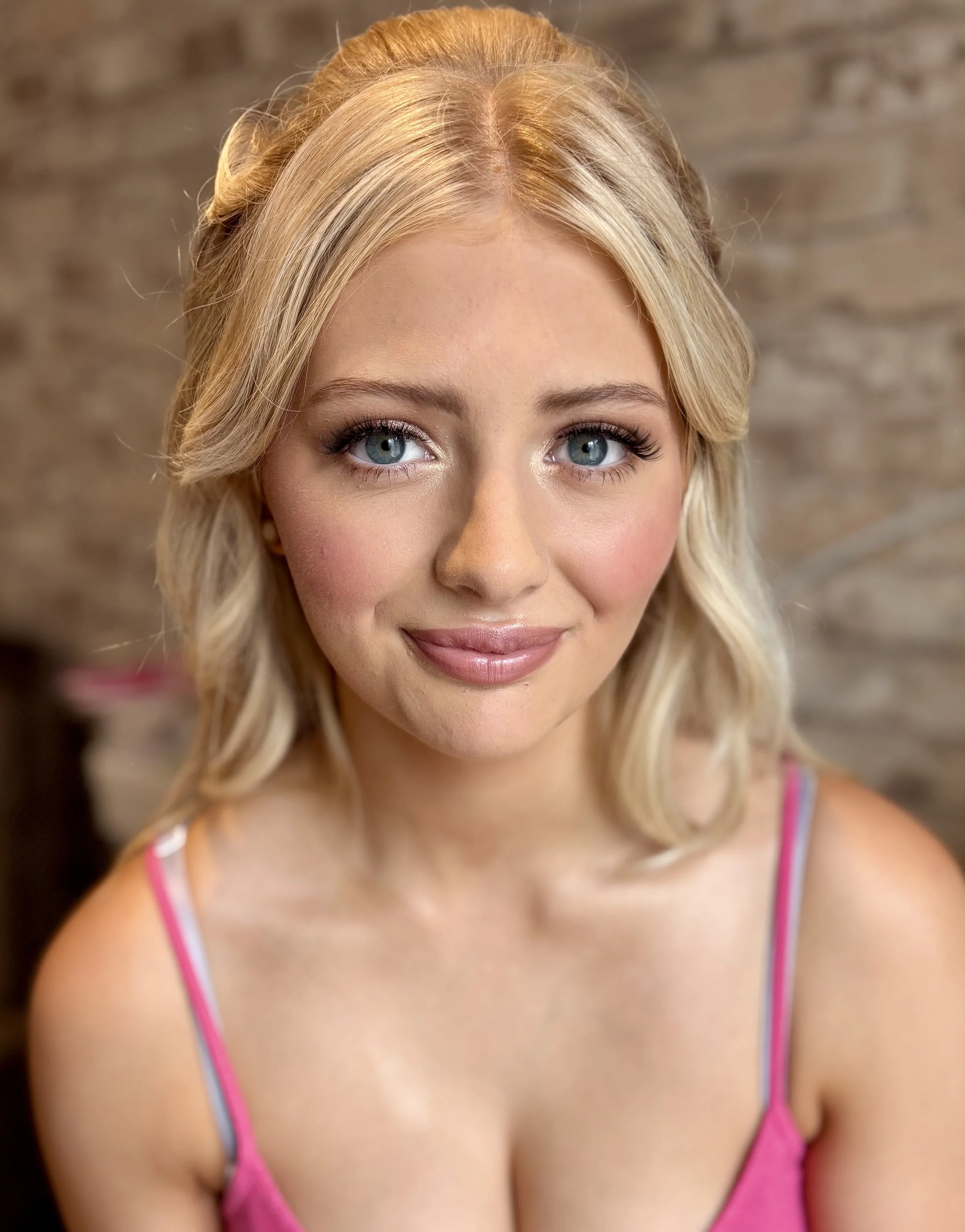 Close-up of a young woman with blonde hair and blue eyes, wearing a pink tank top, smiling softly, with a blurred brick wall background.