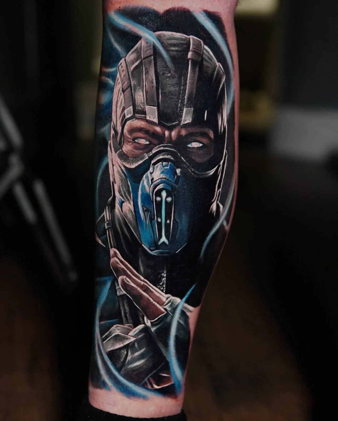 Tattoo of a masked warrior with glowing eyes and crossed arms, featuring blue and black hues, possibly inspired by a video game character.