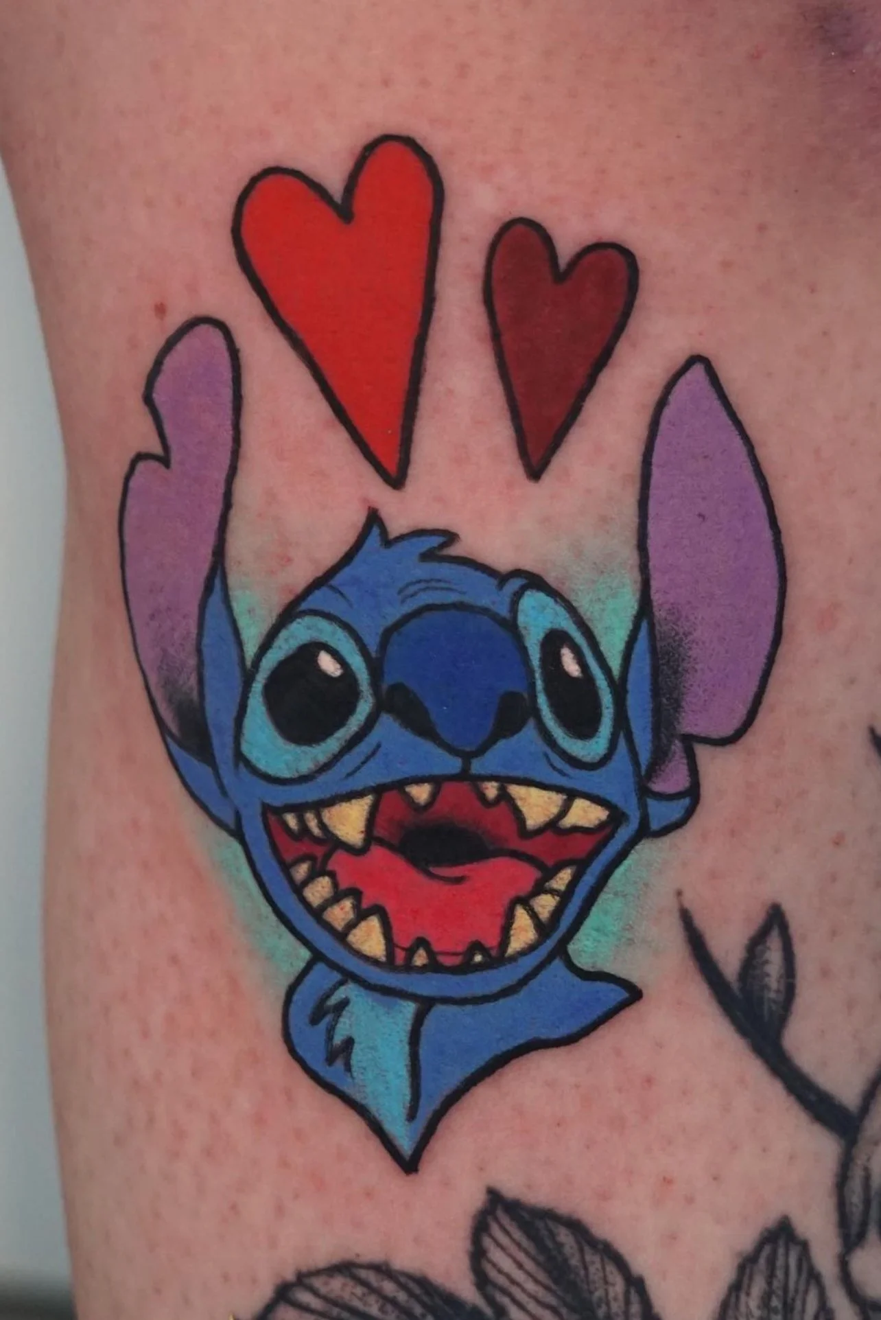 Tattoo of animated blue character with large ears and two red hearts above, on skin with another tattoo nearby.