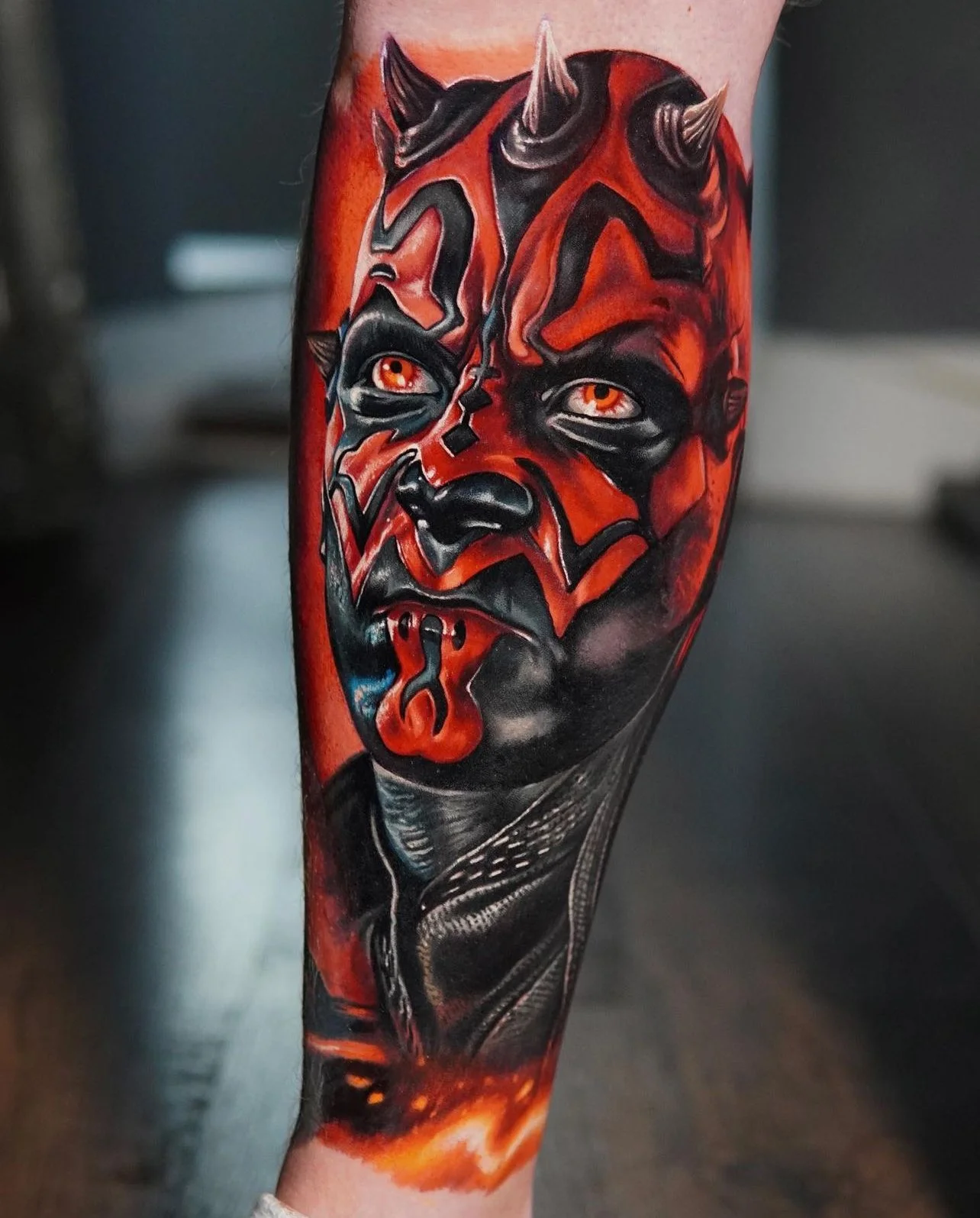 A detailed tattoo of a red and black horned character with orange eyes on someone's leg.