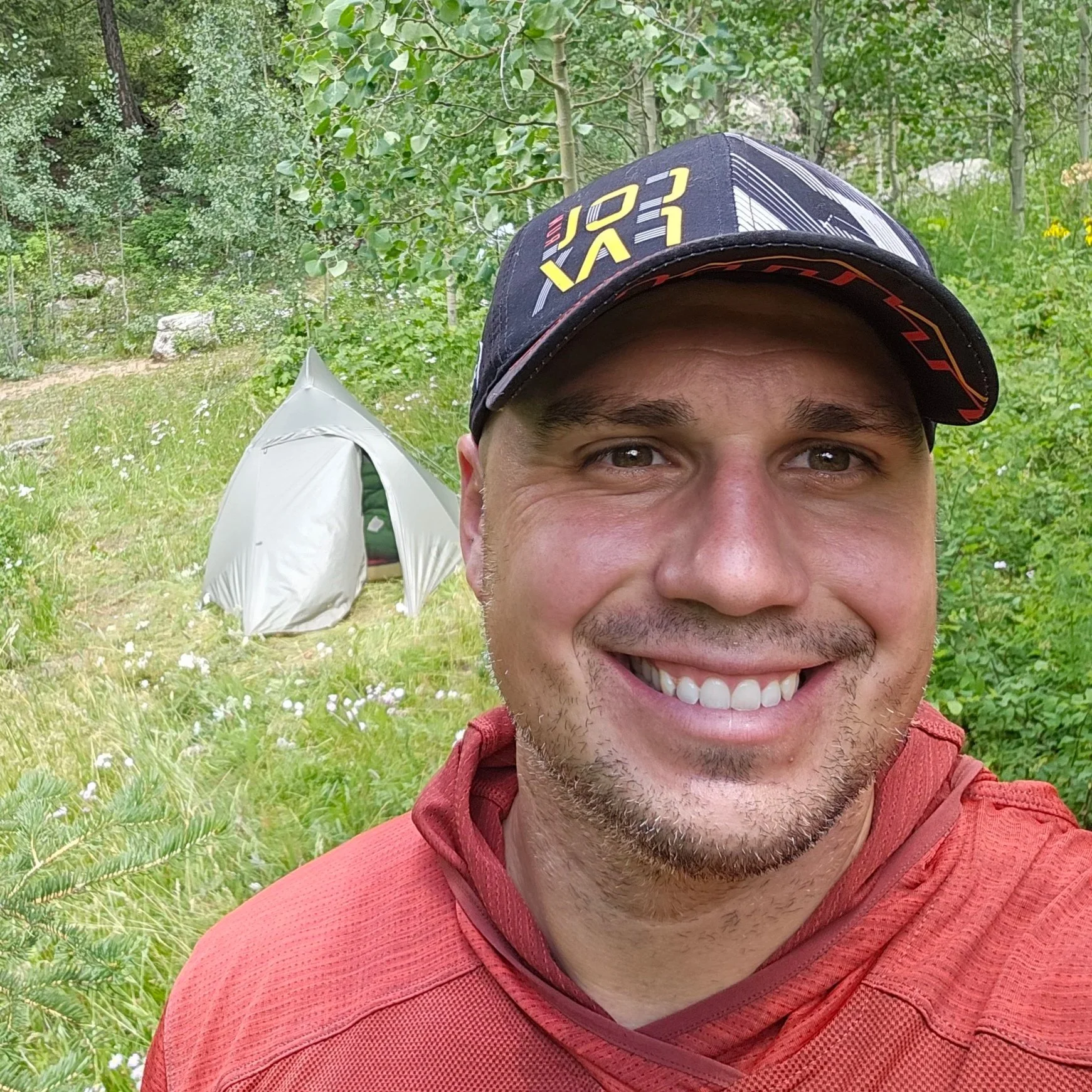 Close-up of a smiling man wearing a black cap and a red outdoor shirt, outdoors with green trees and plants, and a tent in the background.