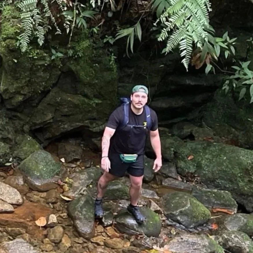 A man hiking in a rocky forested area, wearing black shorts, black shirt, hiking shoes, a green cap backwards, and a small waist pouch.