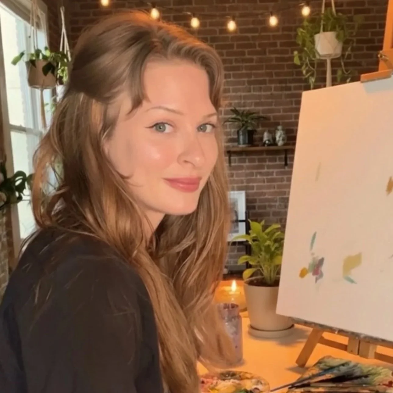 A woman with long, wavy, light brown hair and fair skin is smiling at the camera while painting on a canvas set up on an easel in a cozy indoor space with brick walls, hanging plants, and warm lighting.