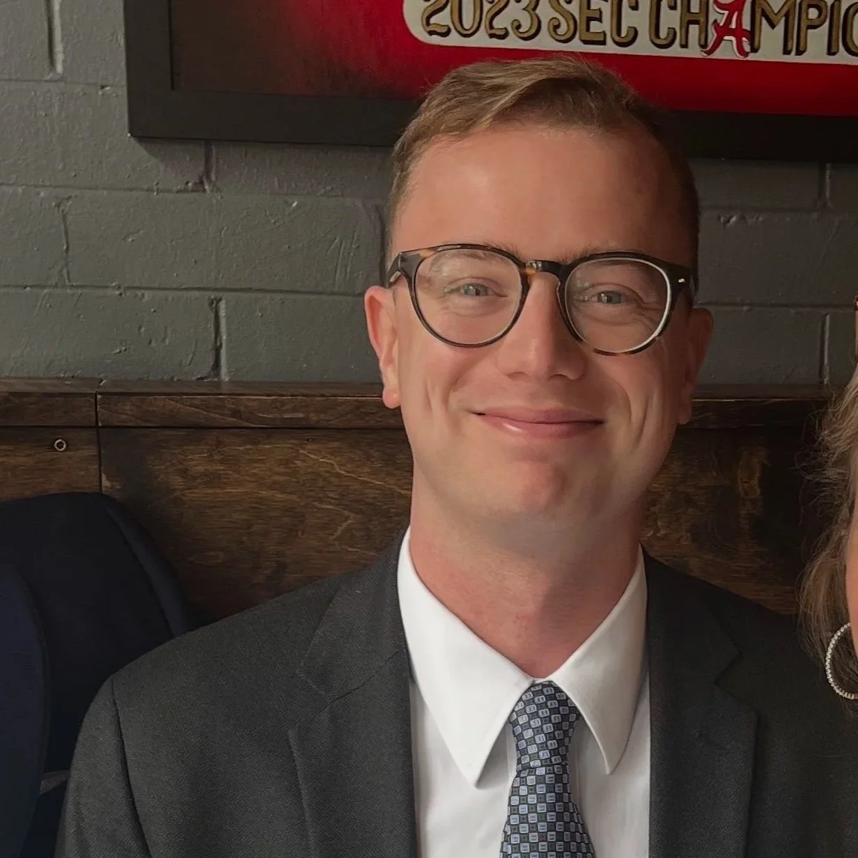 A man in a suit and tie, wearing glasses, smiling, with a gray brick wall and a wooden panel behind him. Part of a sports banner is visible above.