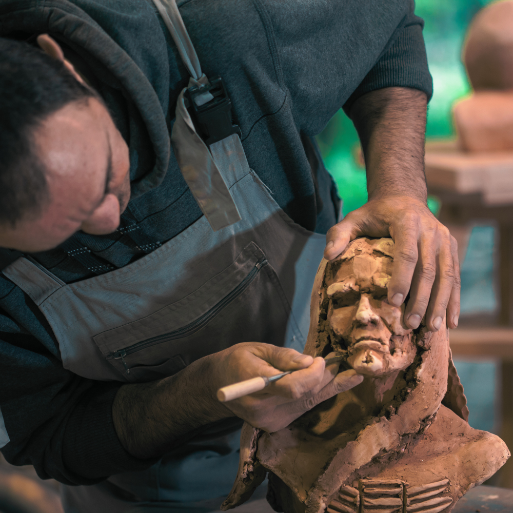 A person working on restoring or sculpting a clay bust sculpture of a man's face and upper torso in an art studio.