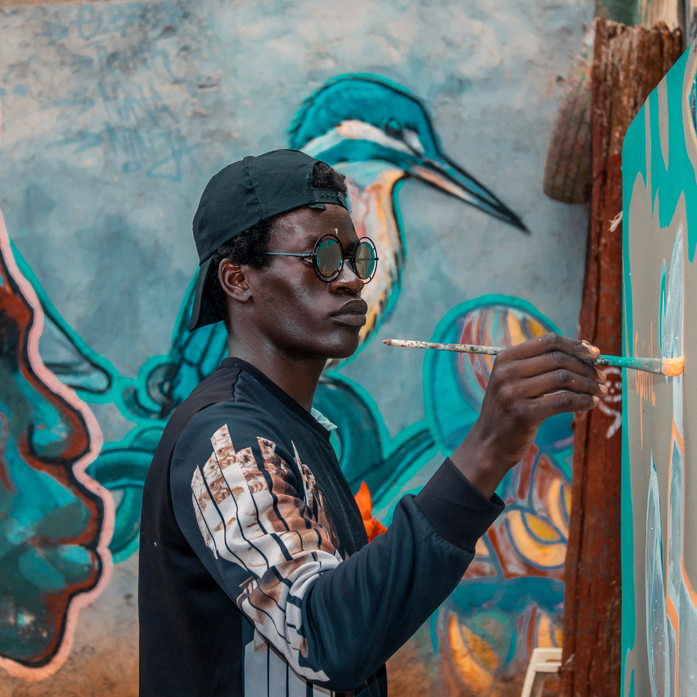 Young Black male artist wearing sunglasses and a backwards baseball cap, painting a colorful mural of a bird on a wall.