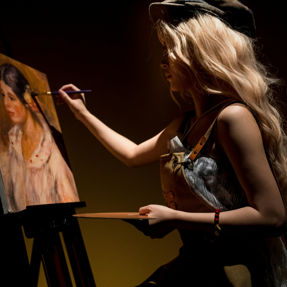 A woman with long blonde hair wearing a black hat and striped apron is painting a portrait on an easel in a dimly lit studio.