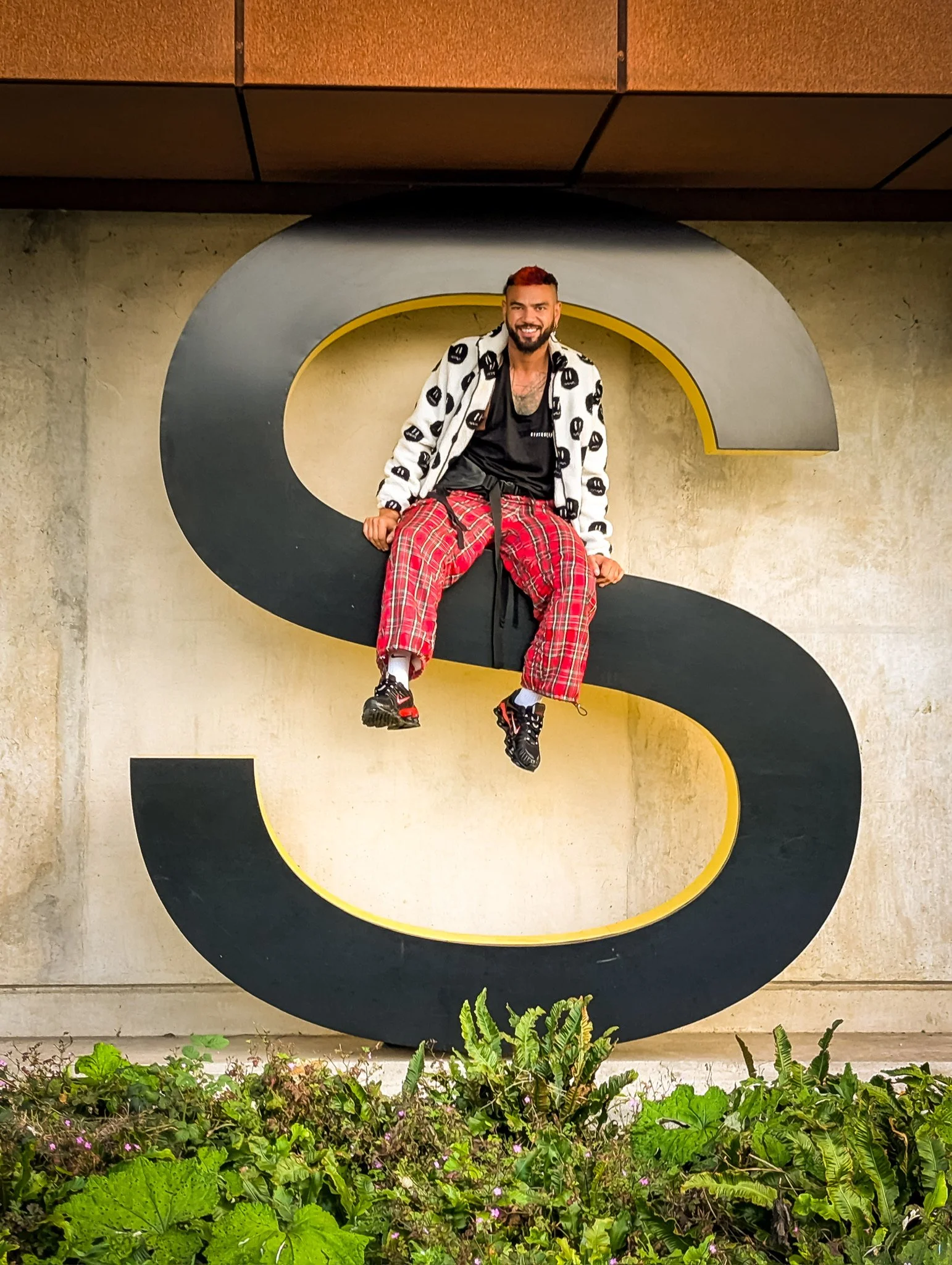 a smiling man sitting on a large decorative letter S in front of a concrete wall with a large black and yellow letter S and some green plants at the bottom of the image