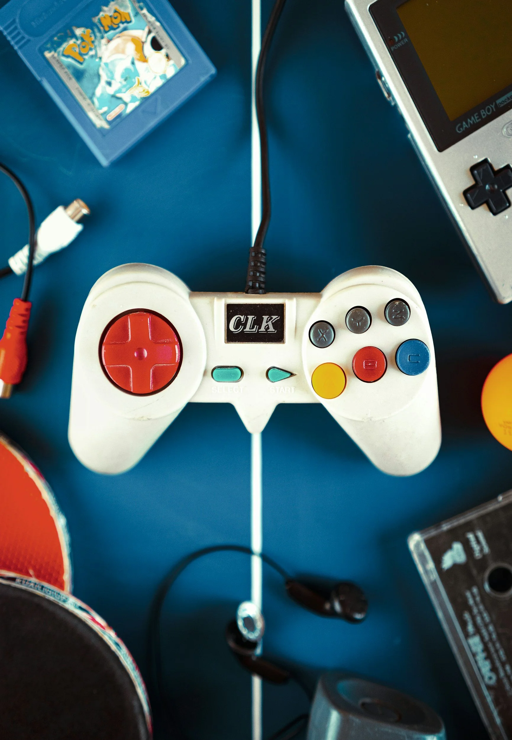 A collection of items on a blue surface including a white game controller with colorful buttons, a black power cord, a Game Boy Advance SP, a tube of white cream, a black headset, a small yellow-orange object, and a black rectangular box.