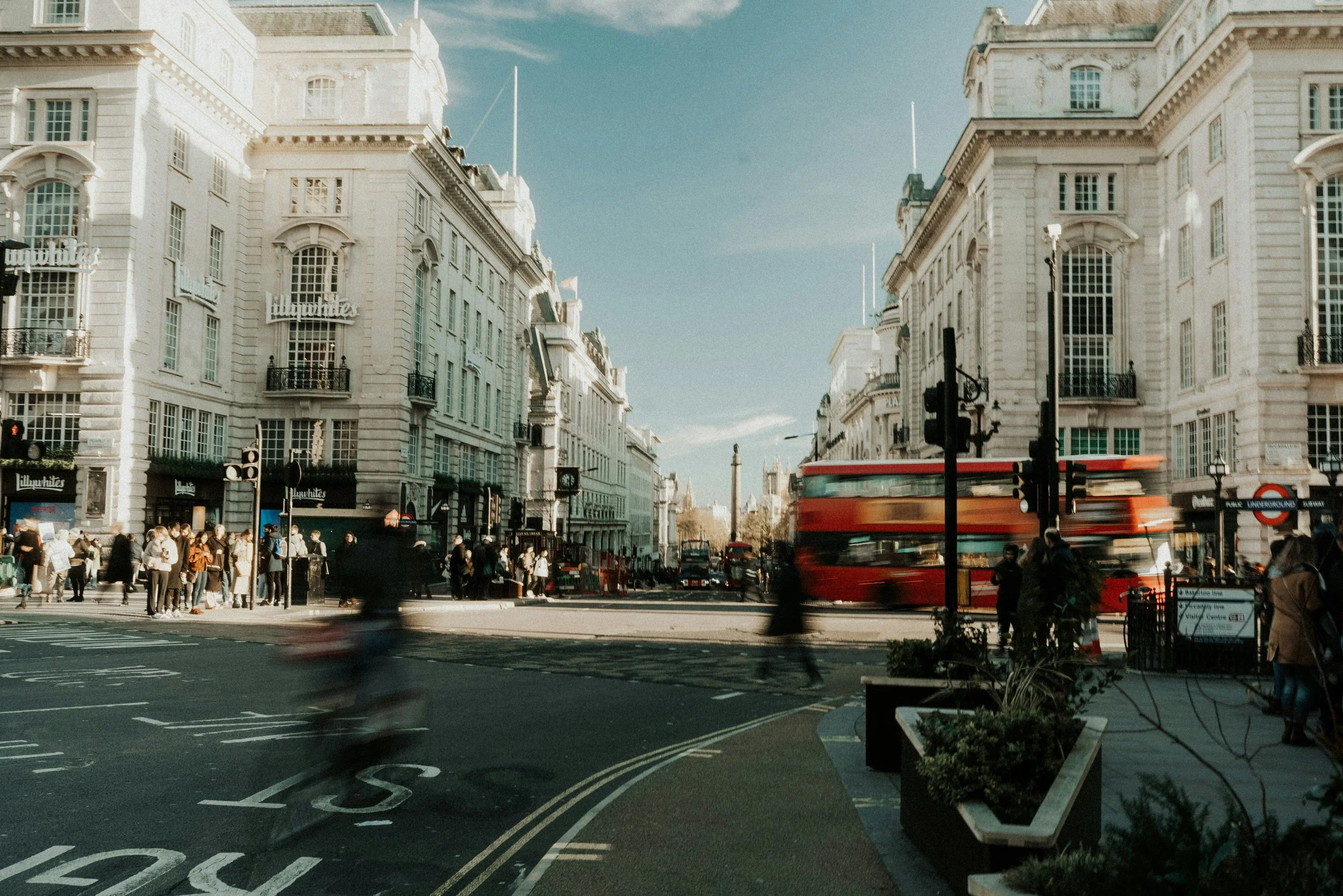 Busy city street in London with large white buildings, a red double-decker bus in motion, and pedestrians crossing the street.