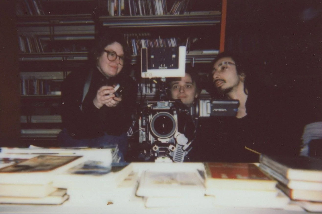 Three men in a bookstore or library taking a selfie with a professional camera, surrounded by books on shelves and a table with books in the foreground.