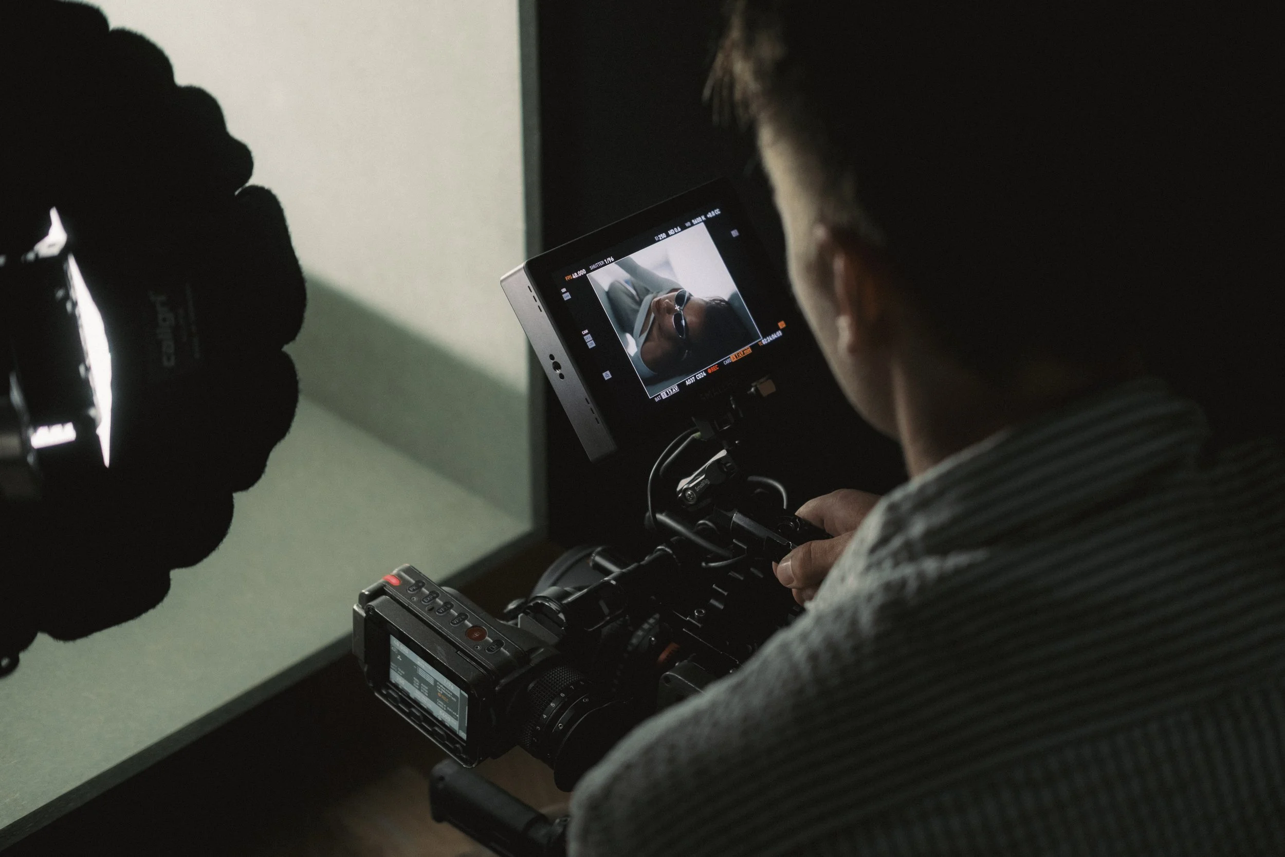 A person operating a professional video camera with a monitor showing a close-up of a person wearing glasses.