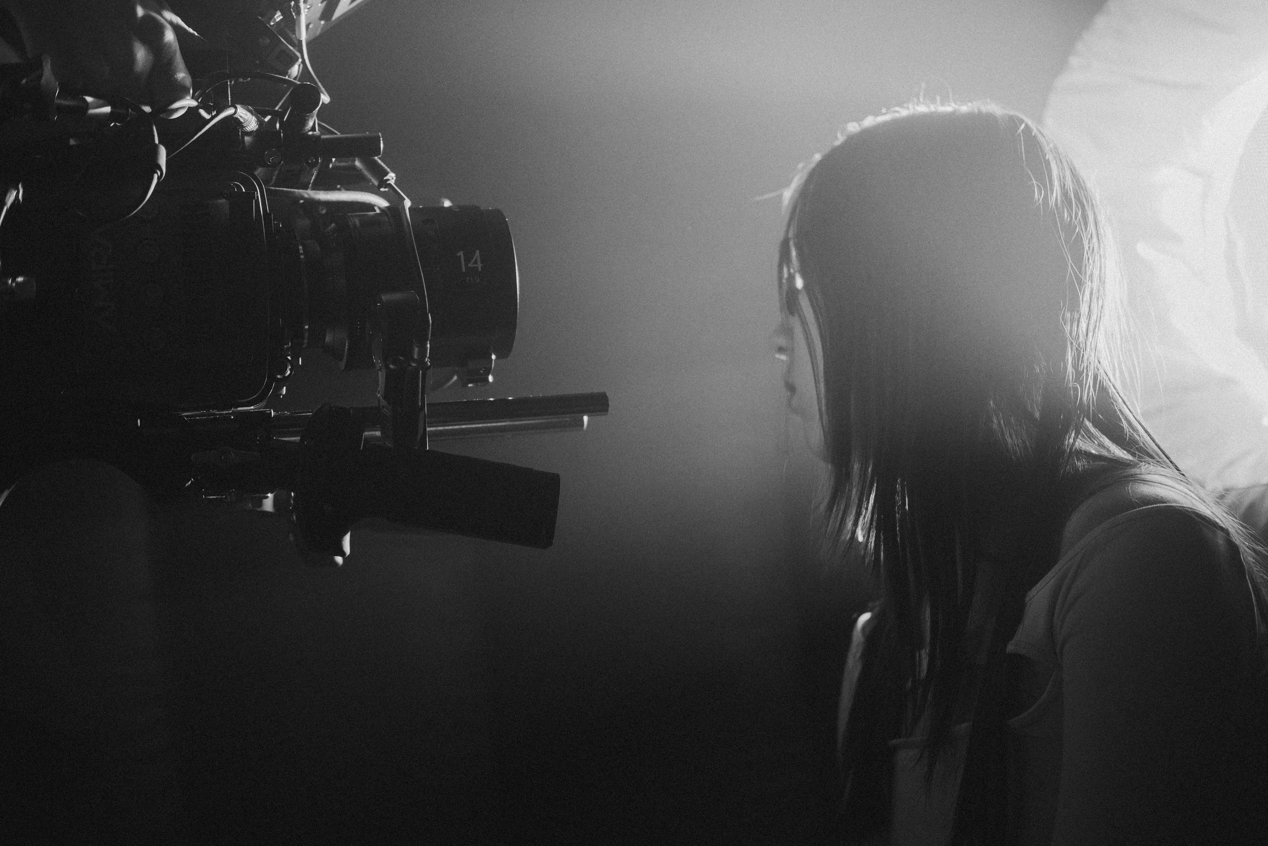 A woman with wet hair facing a large camera with a bright light source behind her in a black-and-white photo.