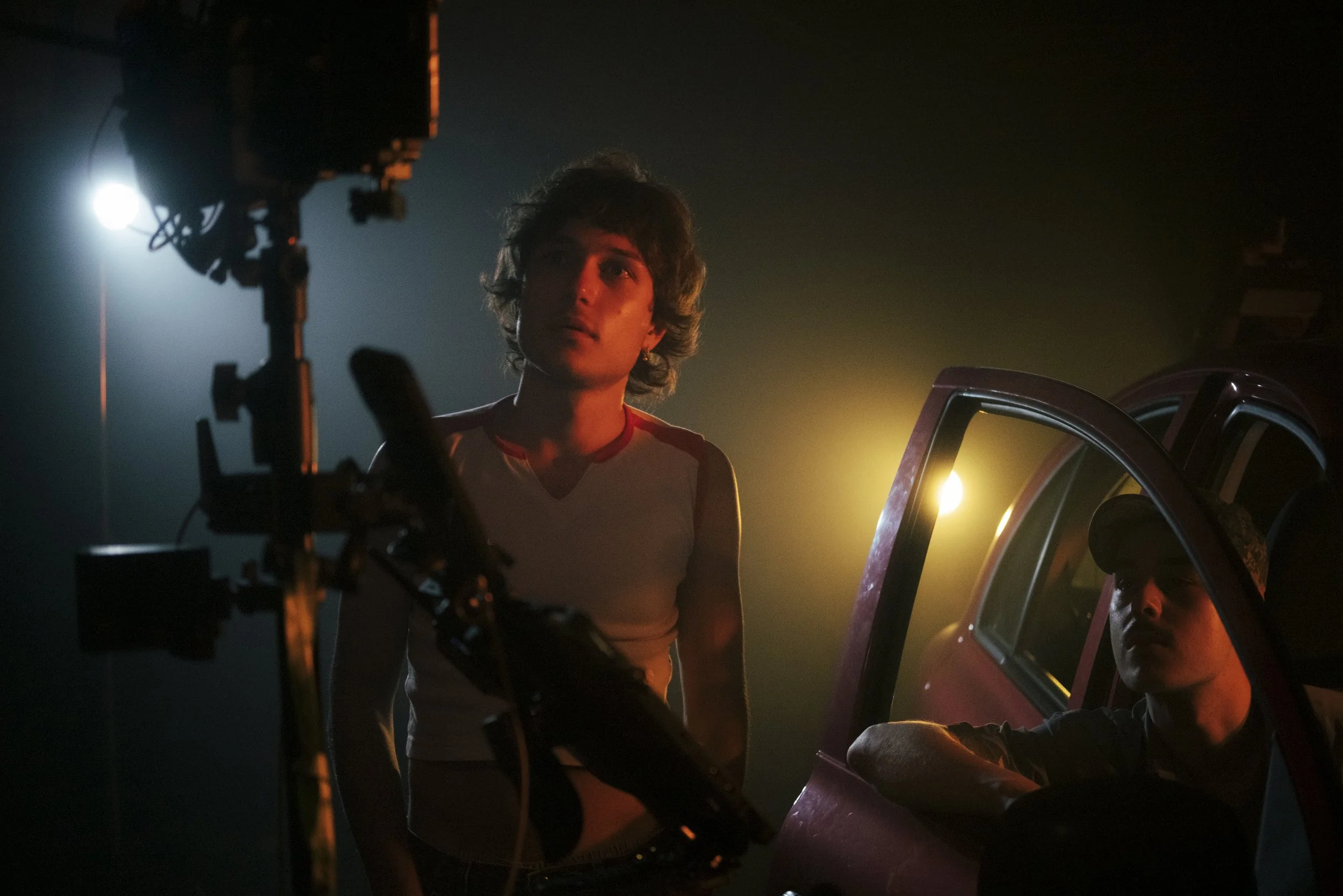 A young man with wavy hair standing next to a man sitting in an open car door at night, with studio lighting illuminating the scene in a dim environment.