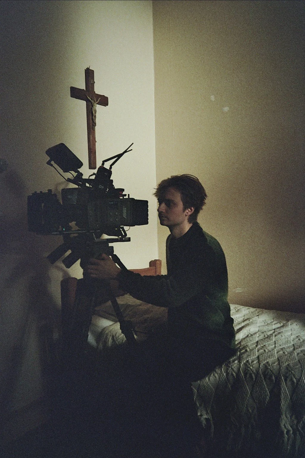 A young man operating a professional video camera in a dimly lit room. There is a wooden crucifix on the wall behind him.