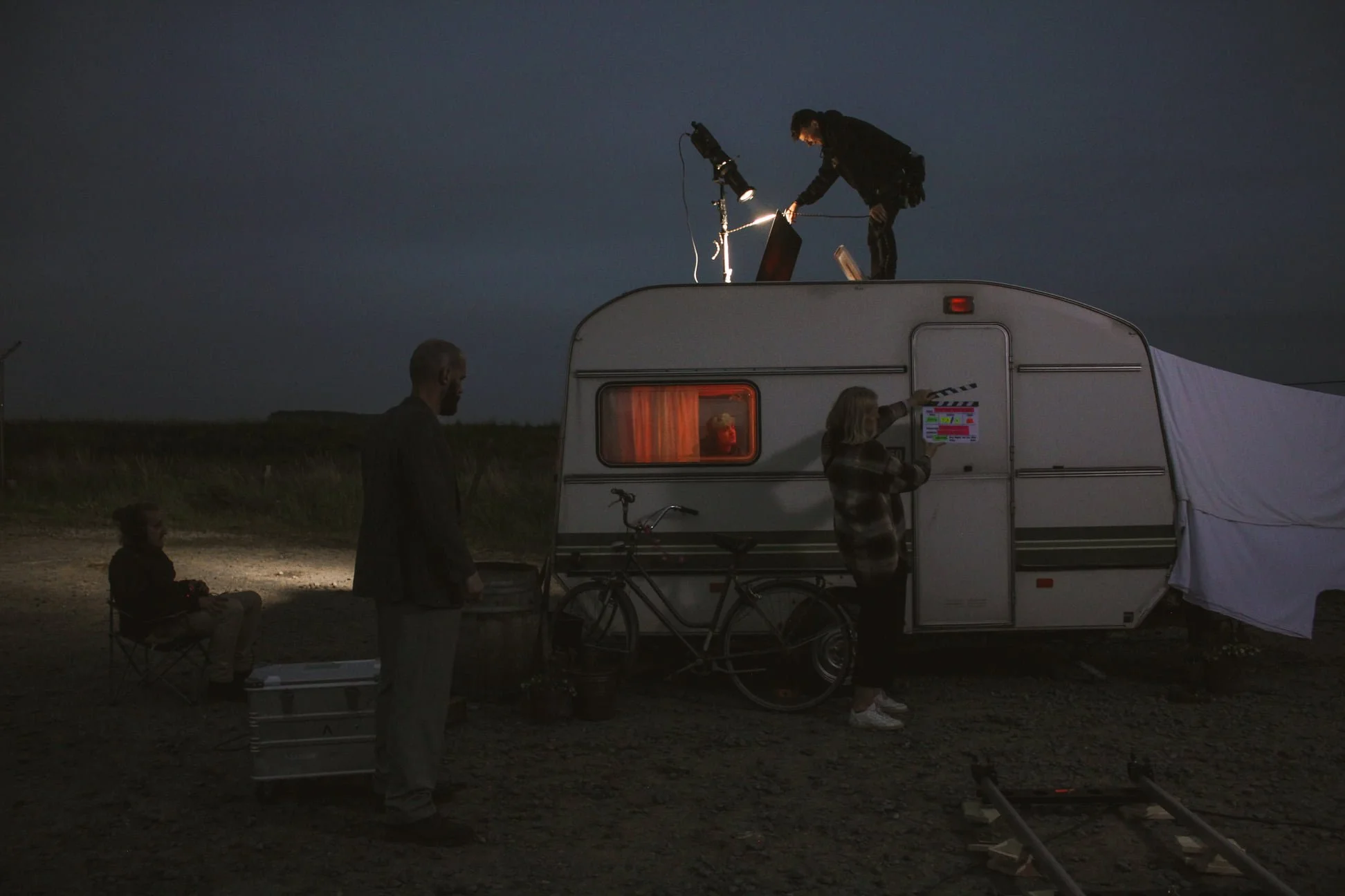 Filming crew working outside a camper at night, with one person on the roof operating filming equipment, and another person operating a clapperboard near the camper, with two more crew members nearby, one seated and one standing.