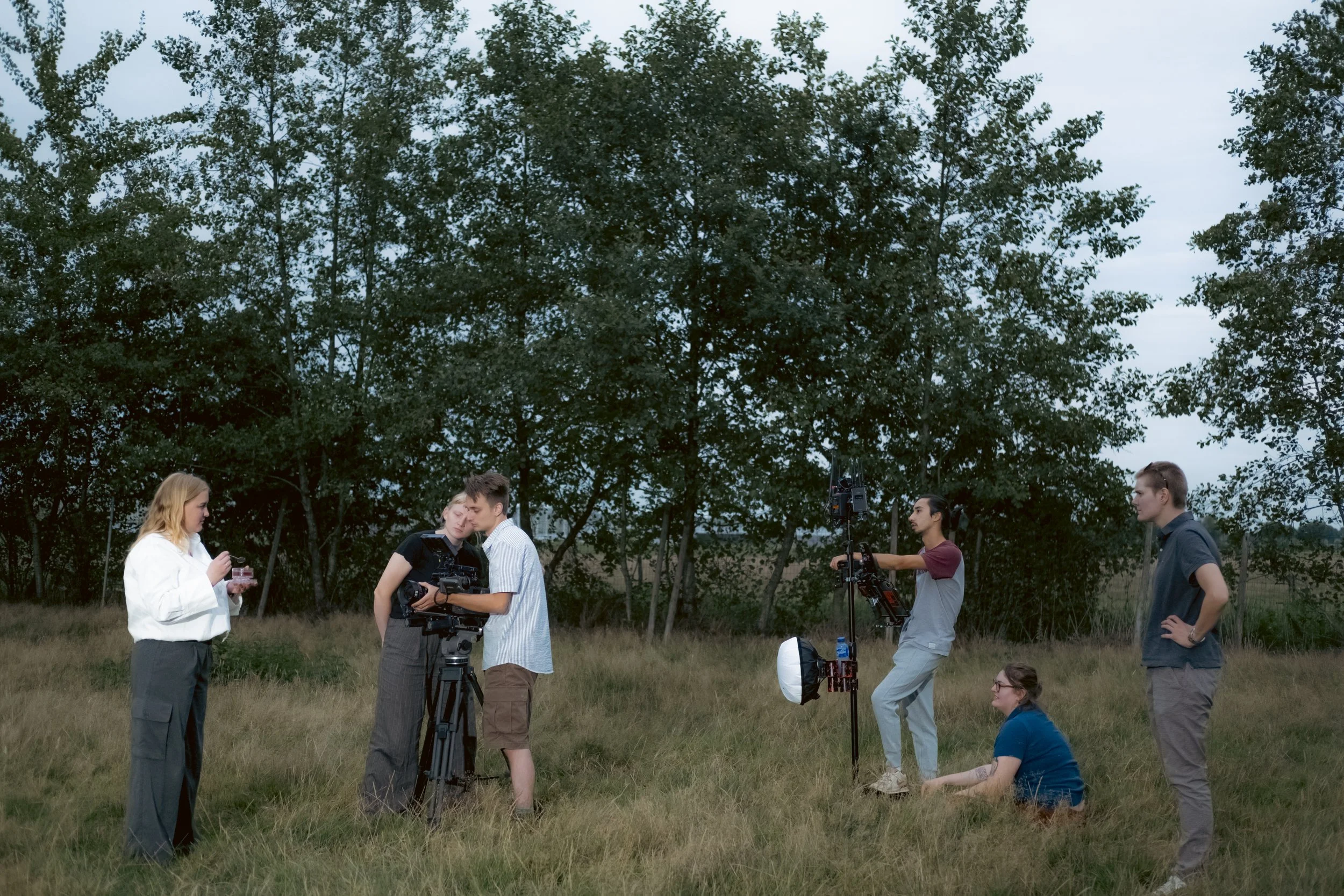 Group of six people working on a film shoot outdoors in a grassy field with trees in the background, some with cameras and equipment.