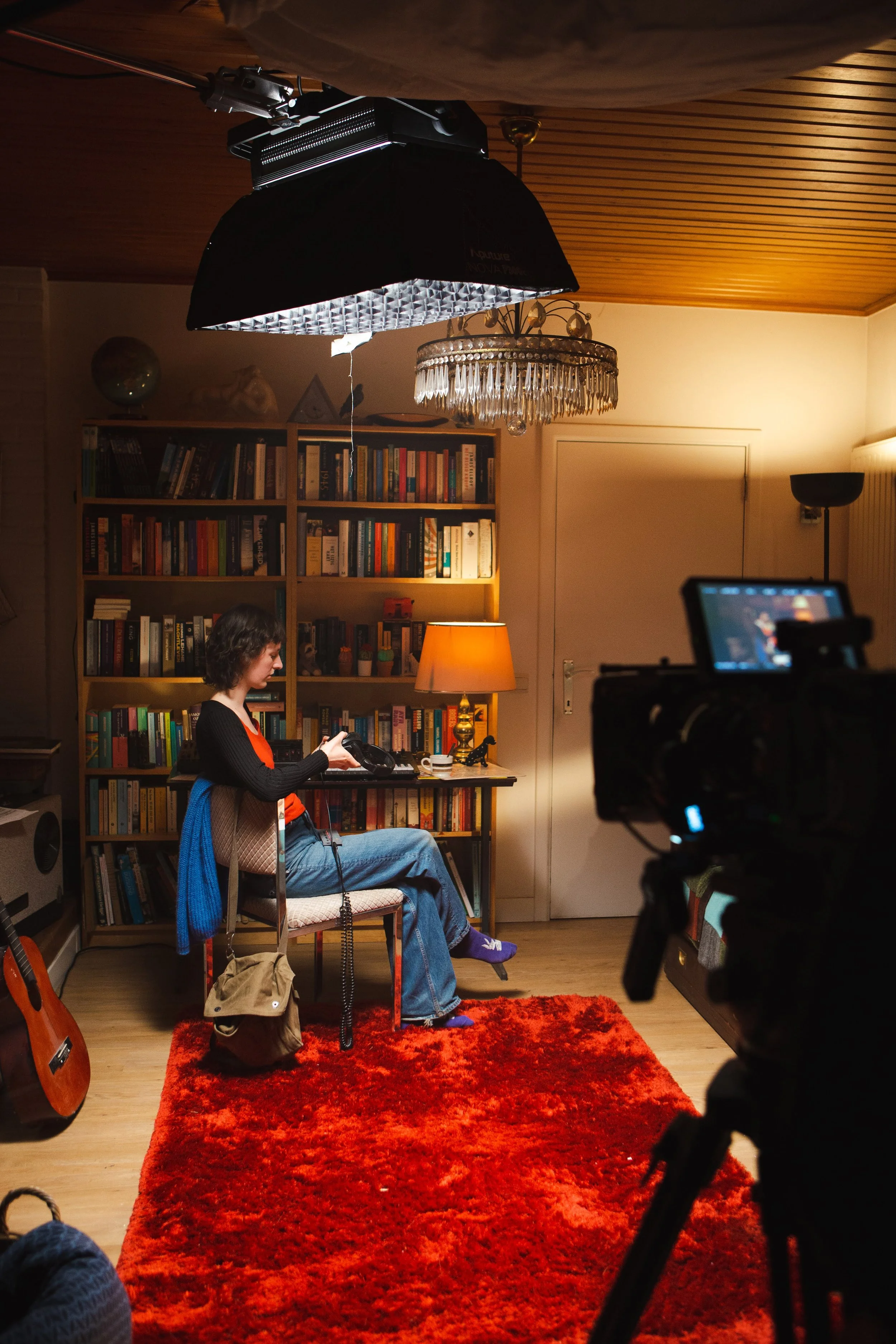 A woman sitting in a room with wooden ceiling and a red carpet, being filmed with professional equipment. She is holding a camera, surrounded by bookshelves, a lamp, a guitar, and various decorations.