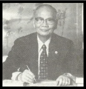 Black and white photo of a man in a suit sitting at a desk with glasses and a pen.