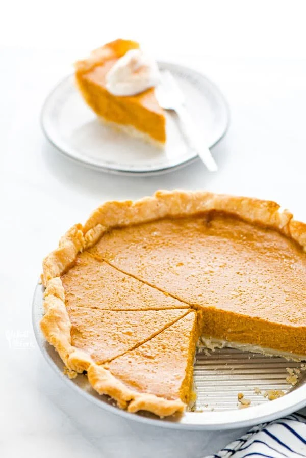 A slice of pumpkin pie with whipped cream on top, placed on a green plate, with a striped orange and green gourd and two green apples in the background.
