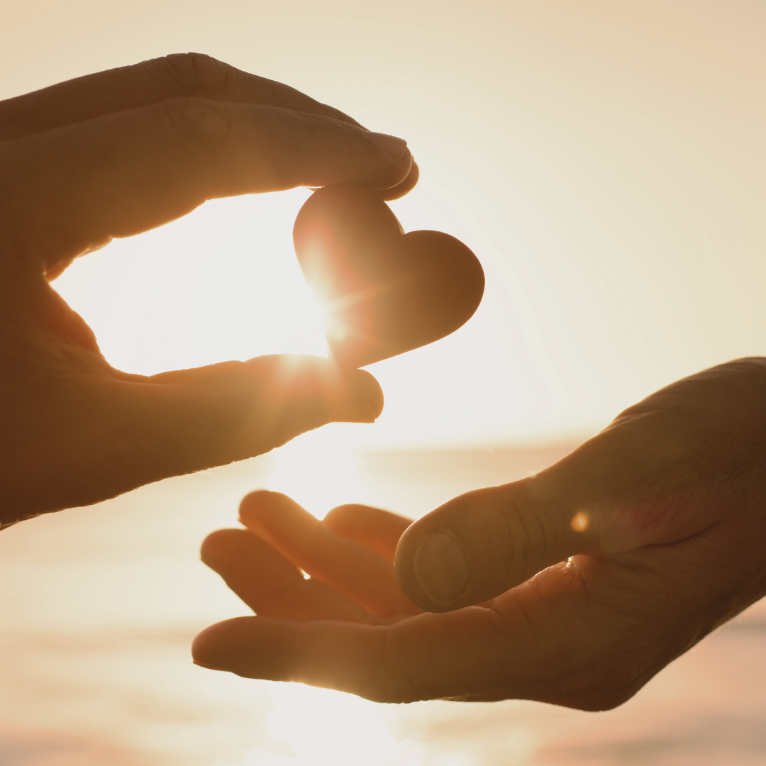 Two hands holding a heart-shaped stone during sunset by the water.